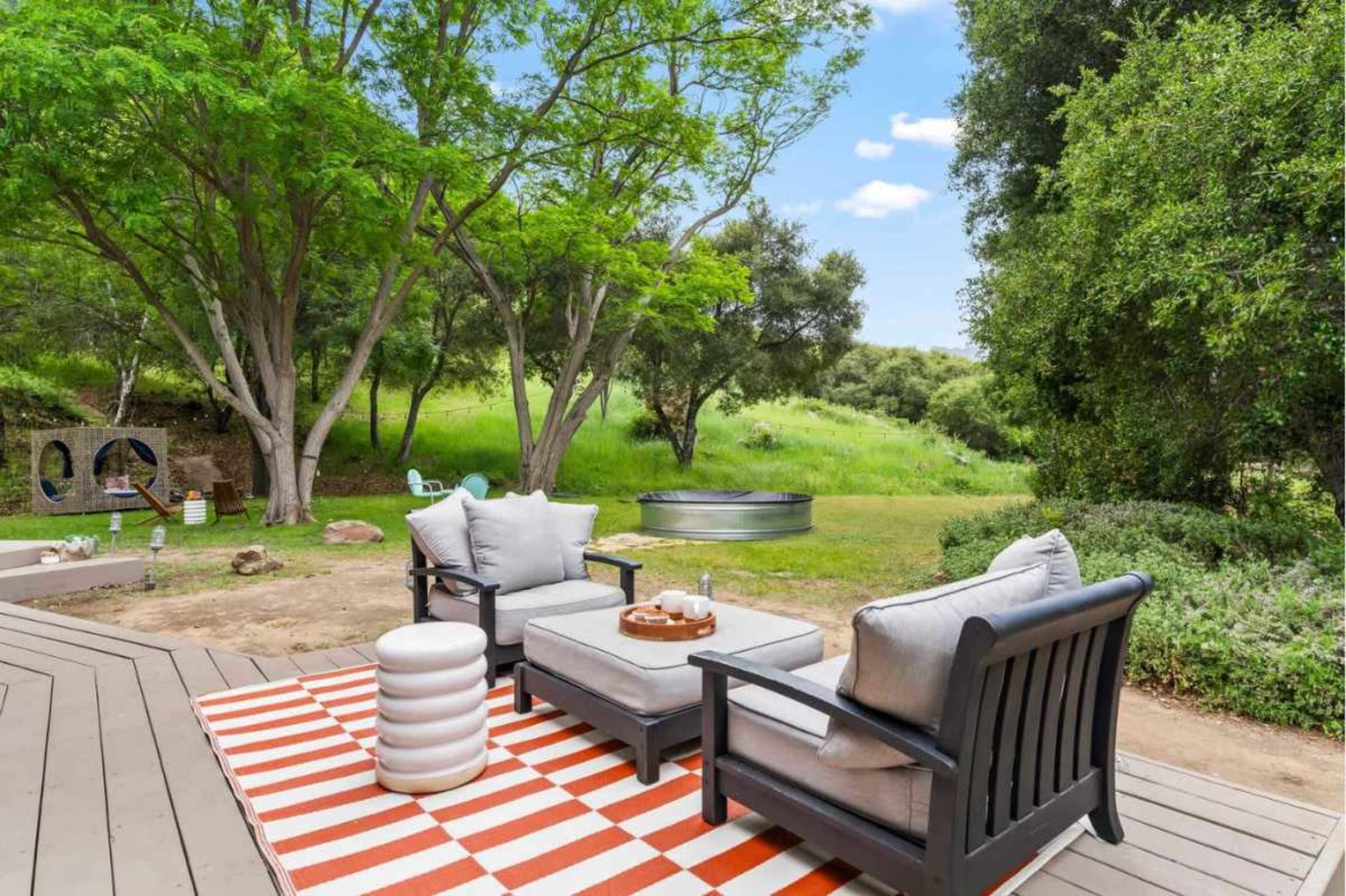 A seating area with two chairs and a small table is set on a deck overlooking a green landscape with trees and a metal trough in the background.