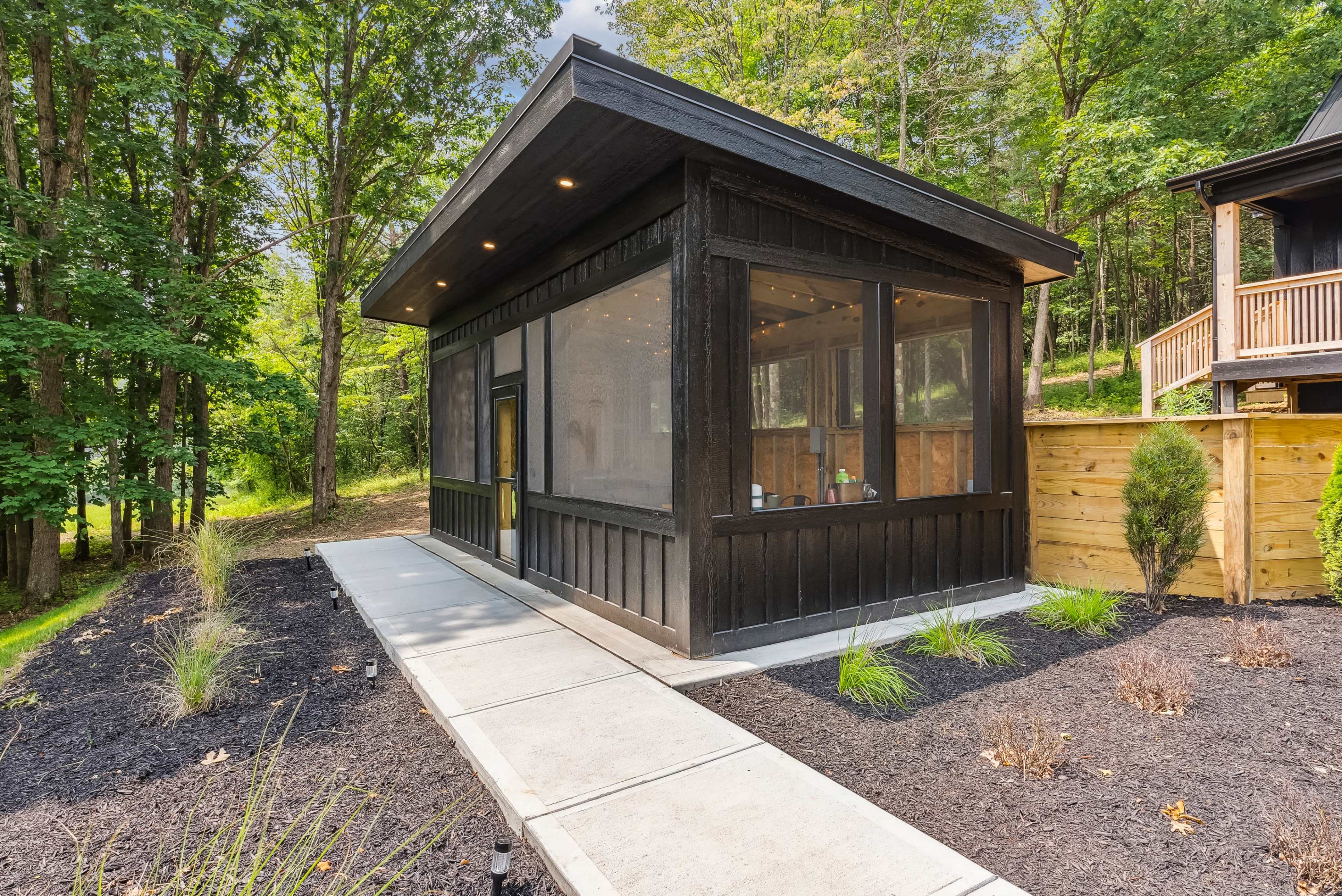 A modern black structure with large screened windows is situated in a wooded area, featuring a concrete path and landscaped planting beds.