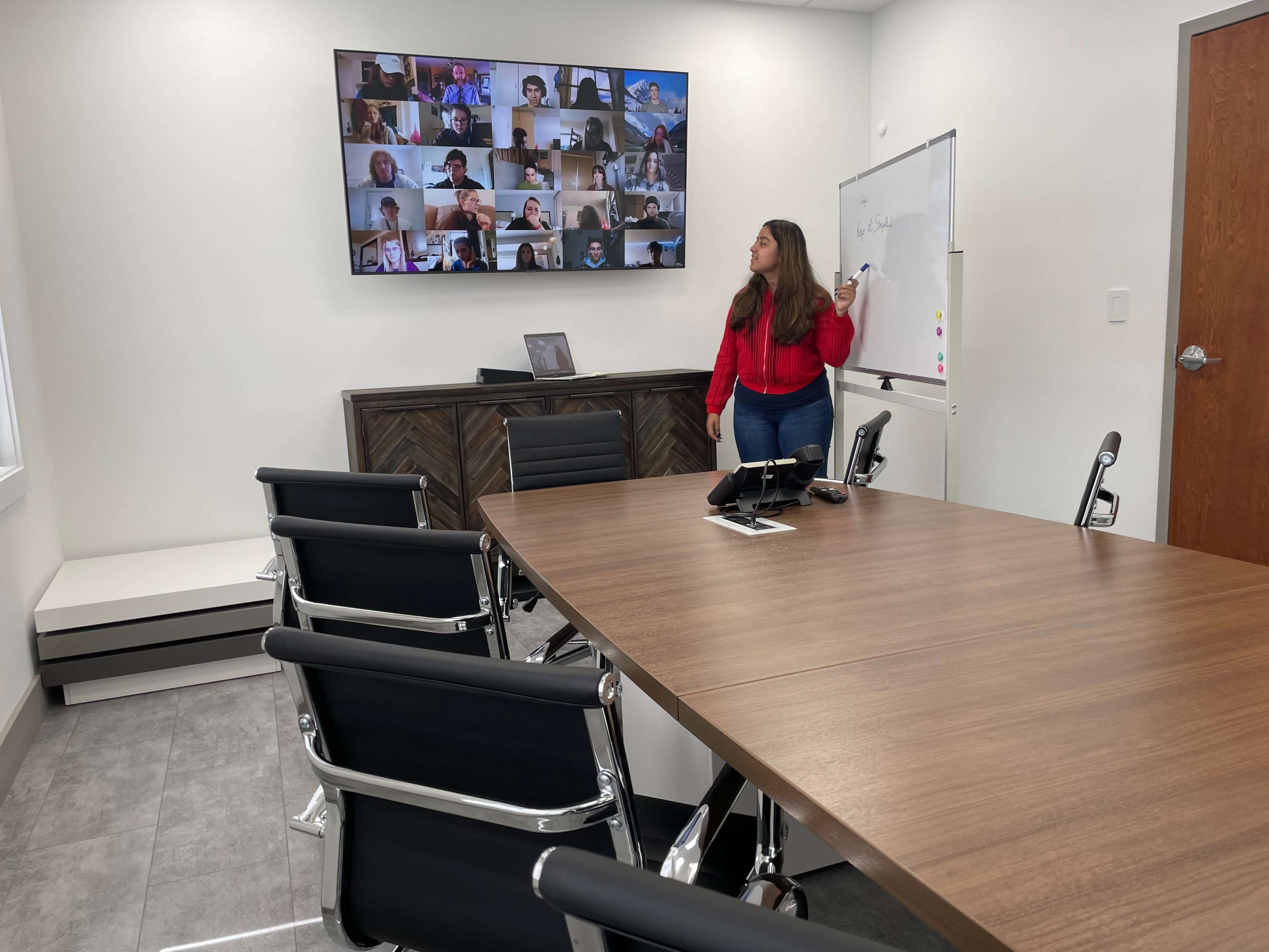 A woman presents in a conference room with a large screen displaying a virtual meeting of multiple participants.
