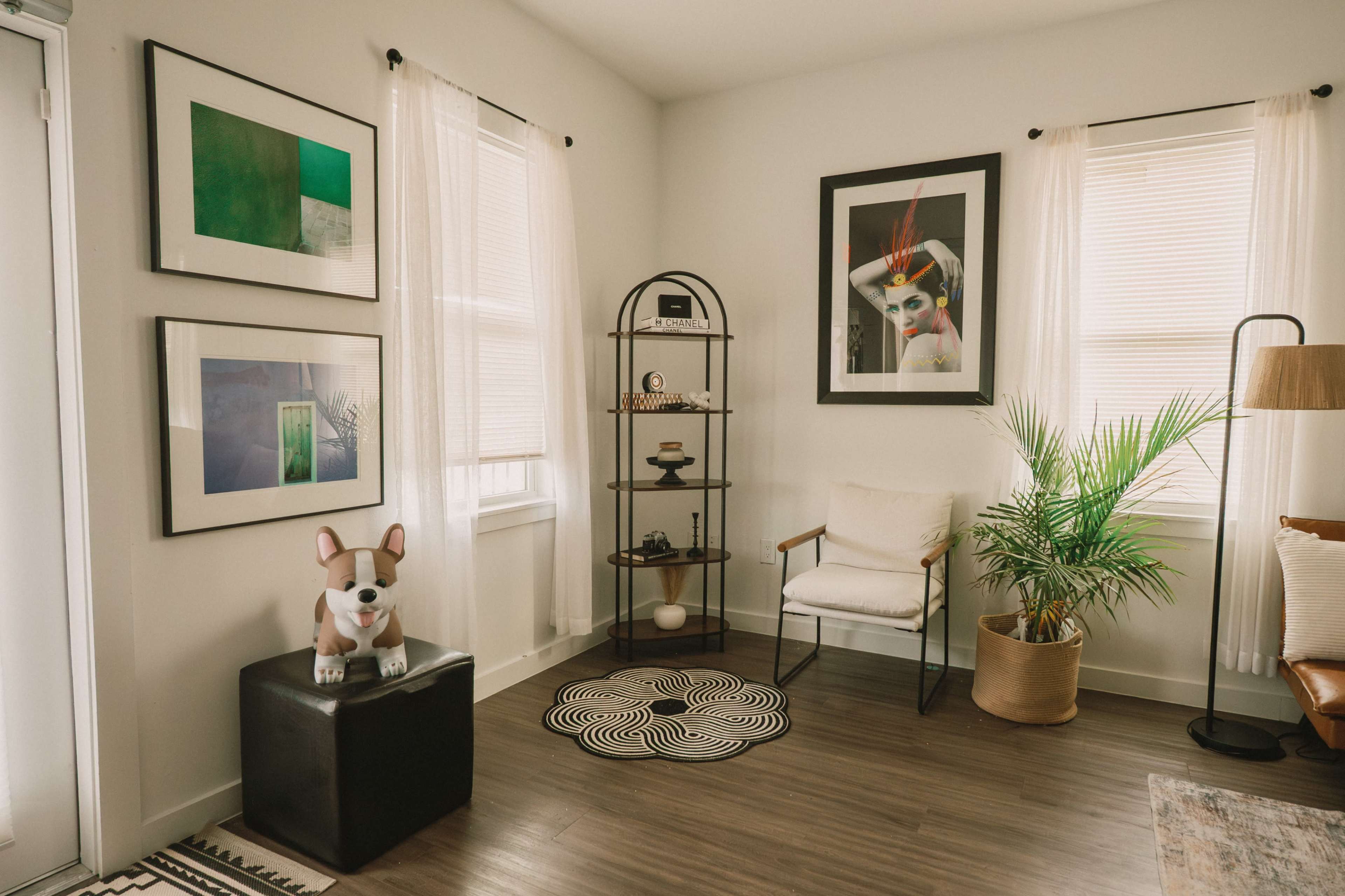 The image shows a cozy interior corner with framed artwork on the walls, a decorative glass shelf, and a small potted plant next to a chair.