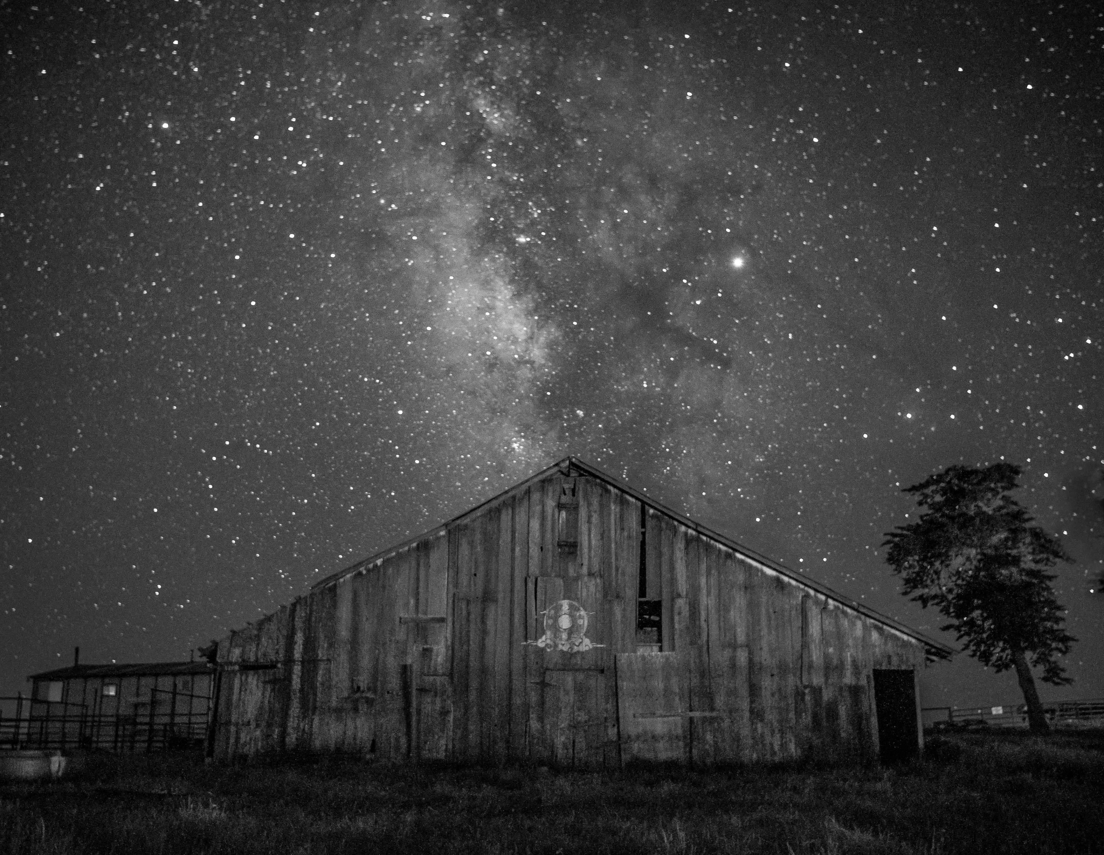 A weathered barn stands under a star-filled sky, with the Milky Way prominently visible above.