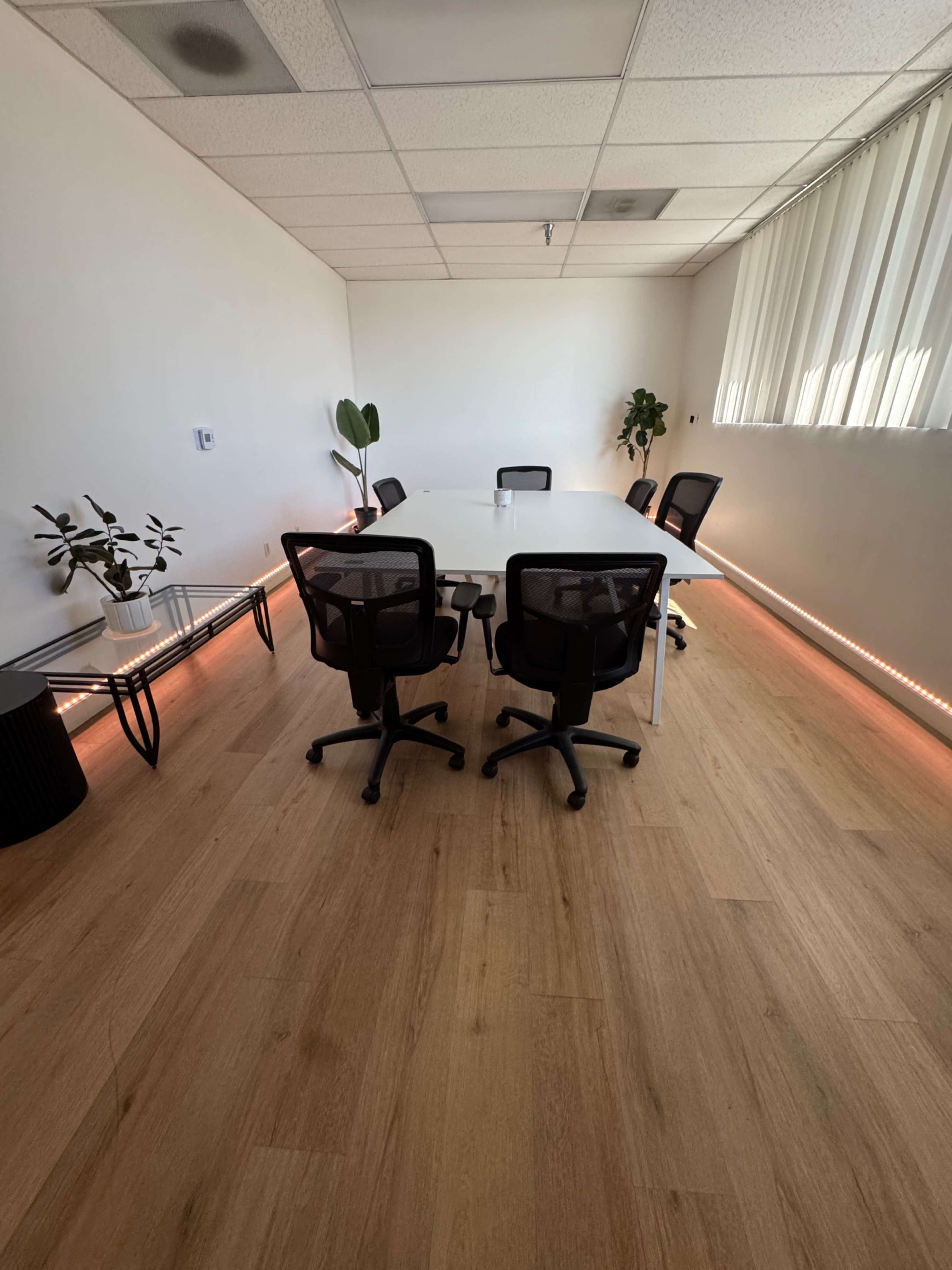 A modern conference room features a white table surrounded by black ergonomic chairs, with plants and ambient lighting along the walls.