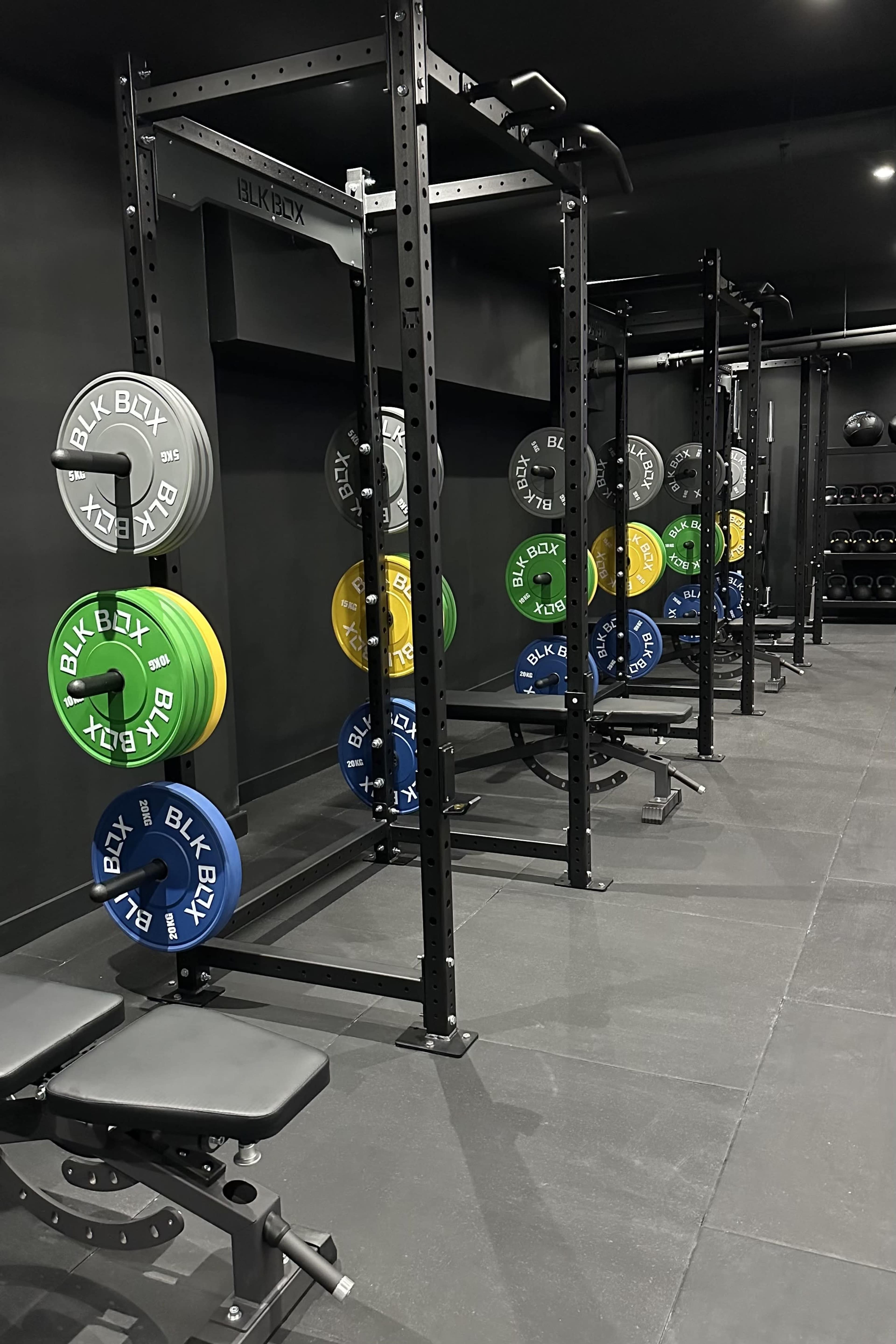 The image shows a well-equipped gym with several squat racks, each loaded with color-coded weight plates, and a bench positioned nearby.