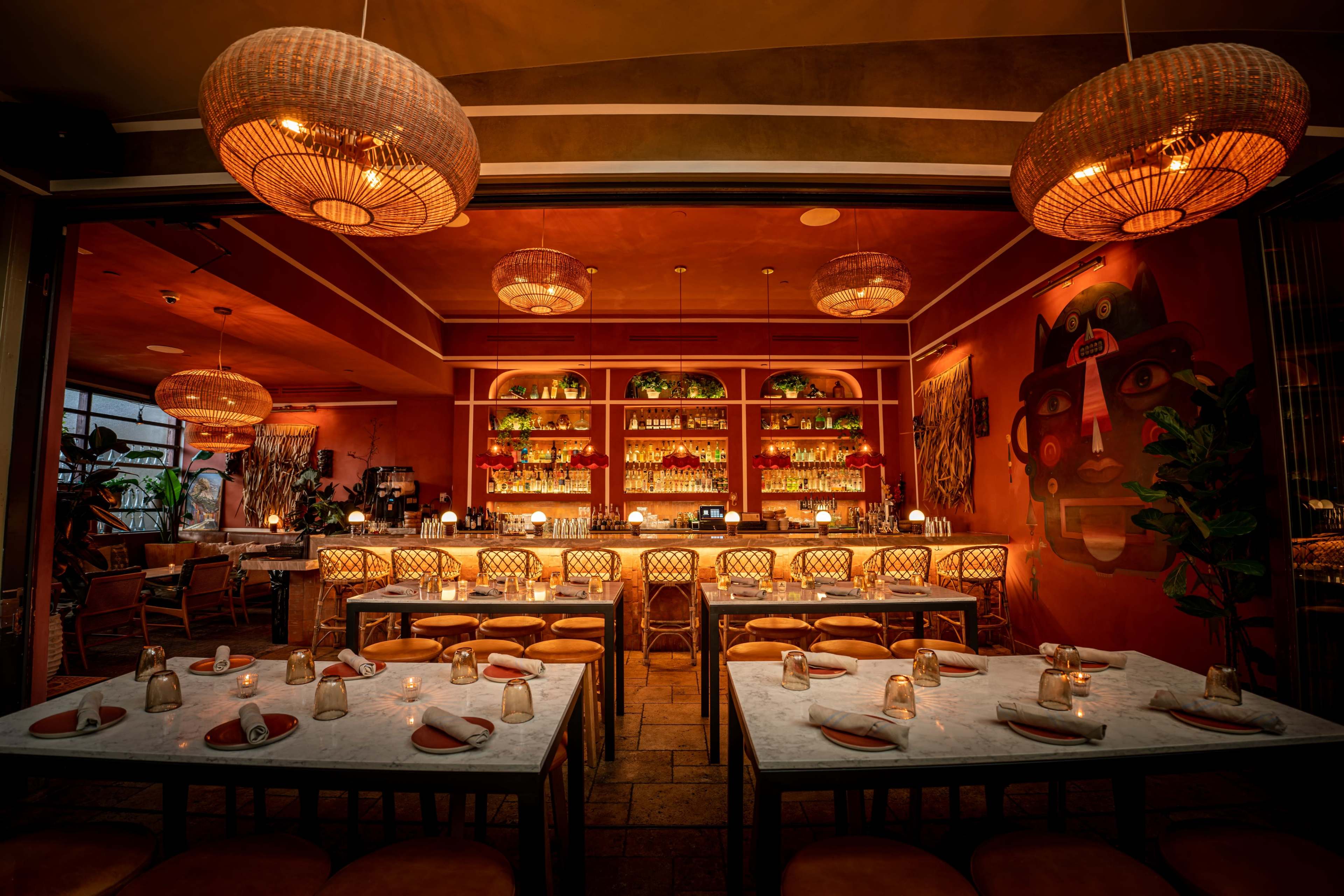 A warmly lit restaurant interior featuring a long bar, rows of tables set for dining, and decorative overhead lighting.