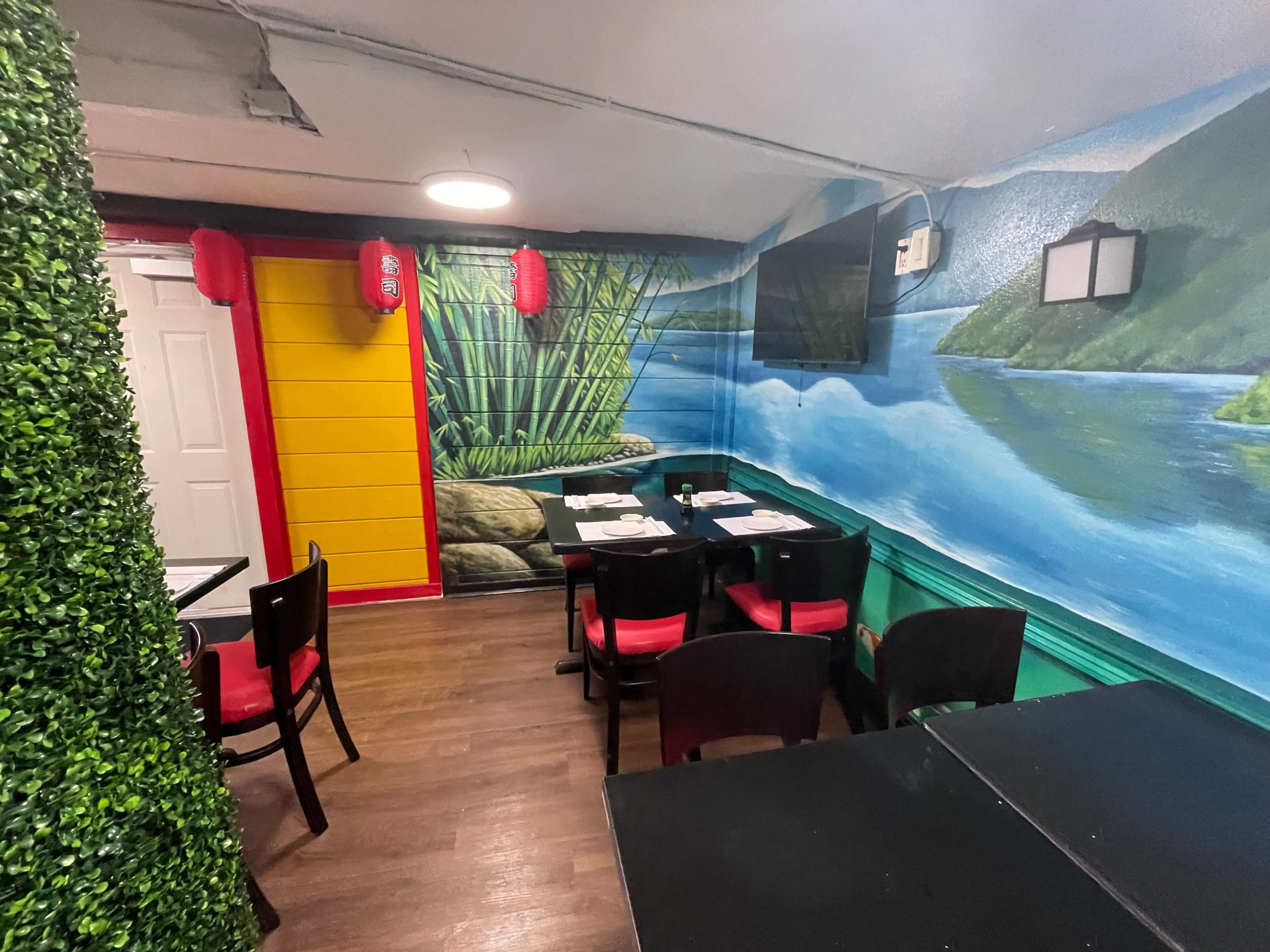 The image shows a restaurant interior featuring colorful murals of a landscape, green plants, and black tables with red and black chairs.