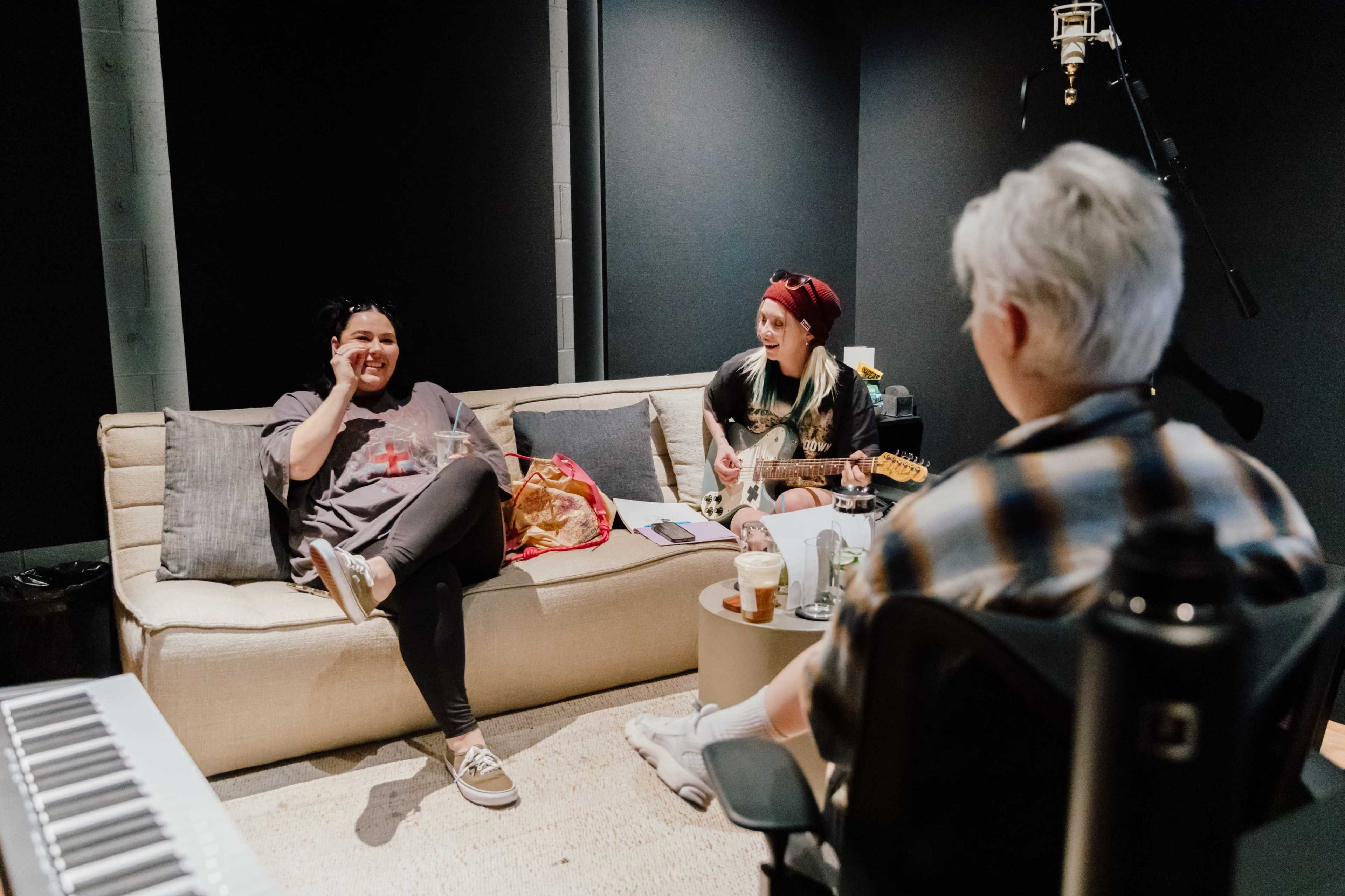 Three people are seated in a recording studio, with one playing guitar, another smiling and holding a phone, and the third focused on them from a chair.