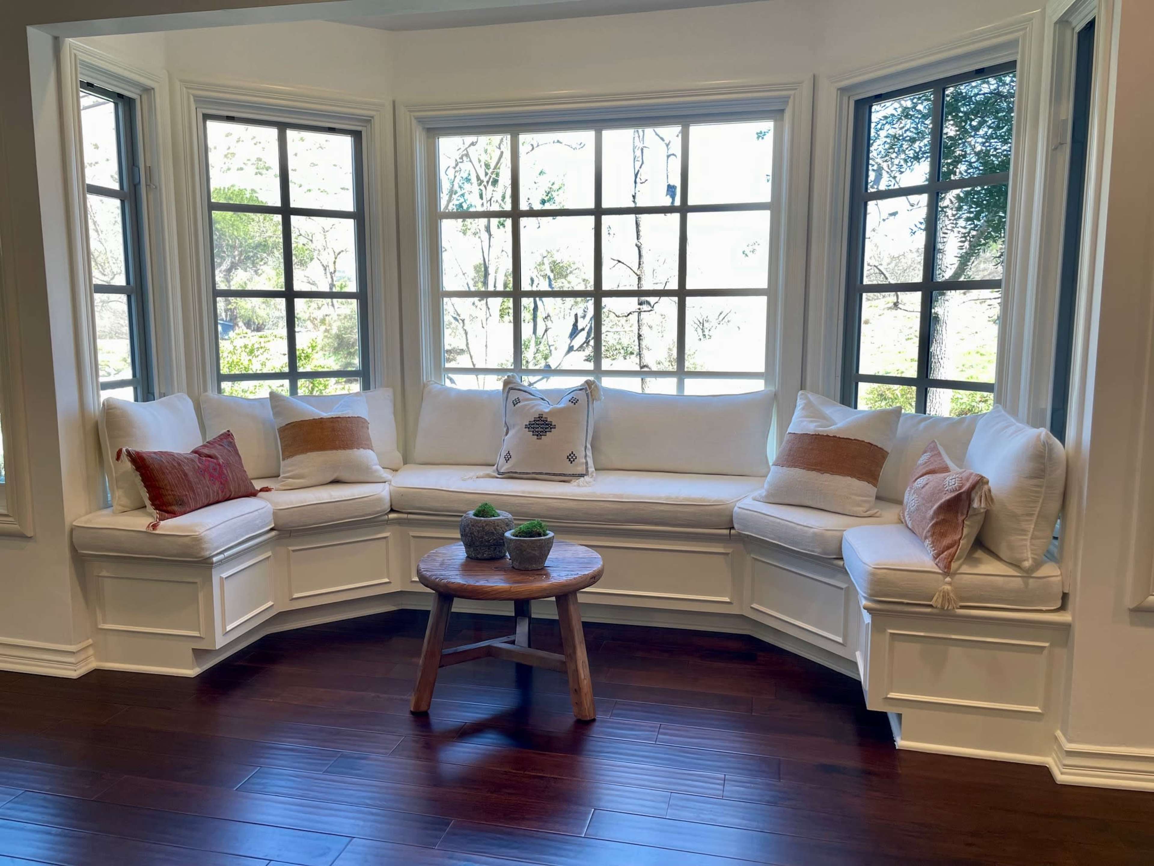 A cozy nook with a white upholstered bench around a round wooden table, surrounded by windows that provide natural light.