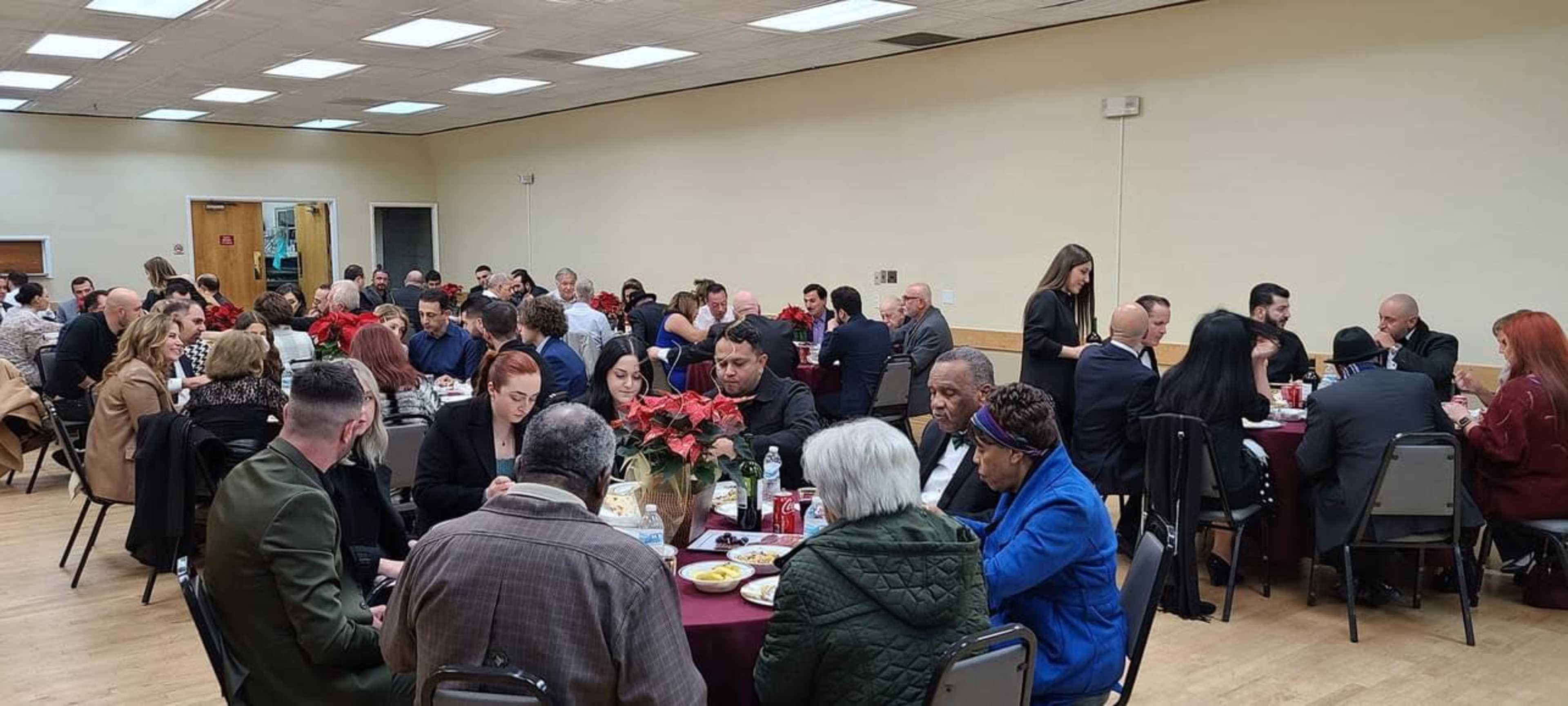 A large group of people sits at tables in a banquet hall, enjoying a meal together.