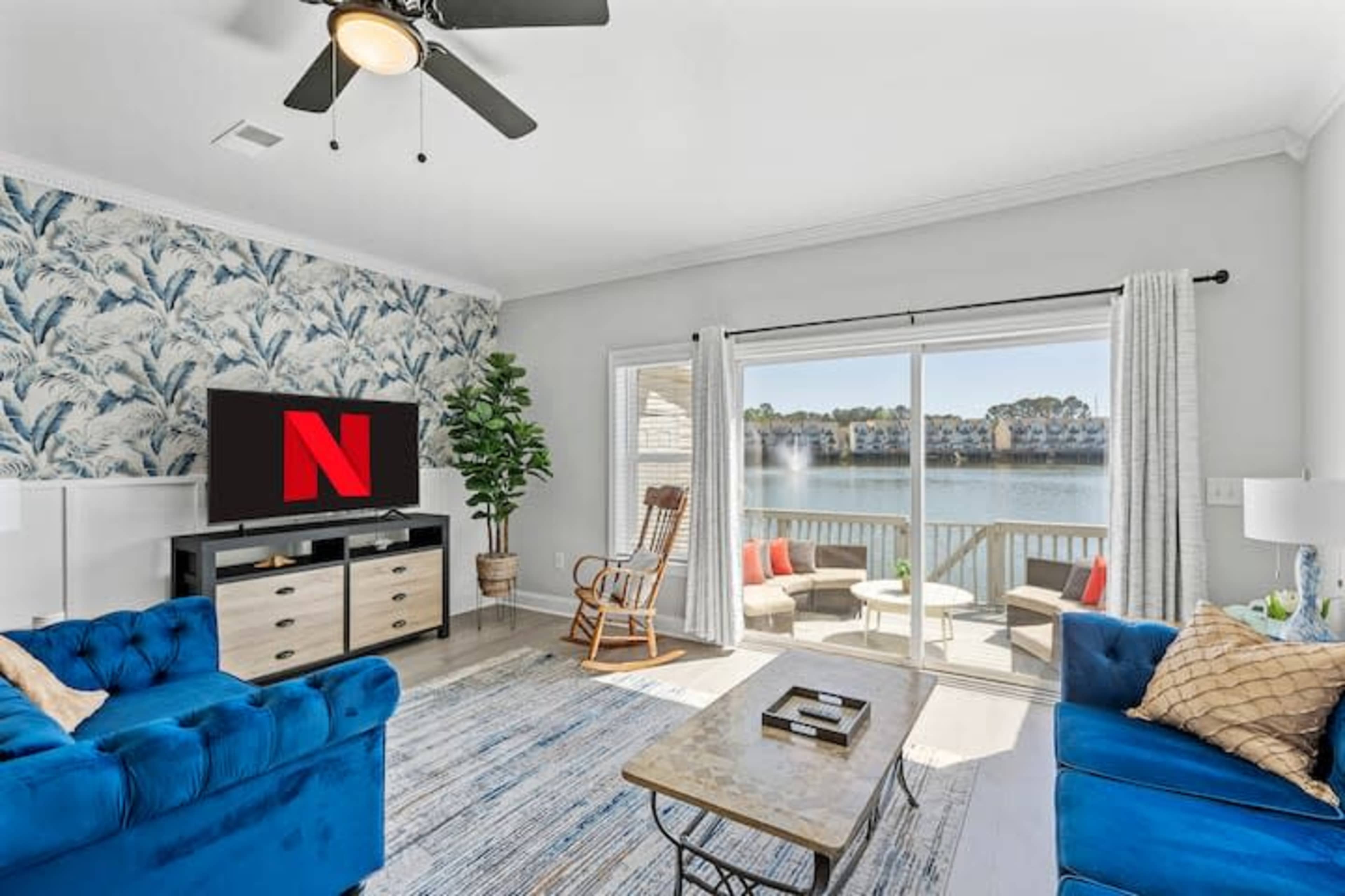 A living room features blue sofas and a wall with tropical wallpaper, with a view of a lake visible through a sliding glass door.