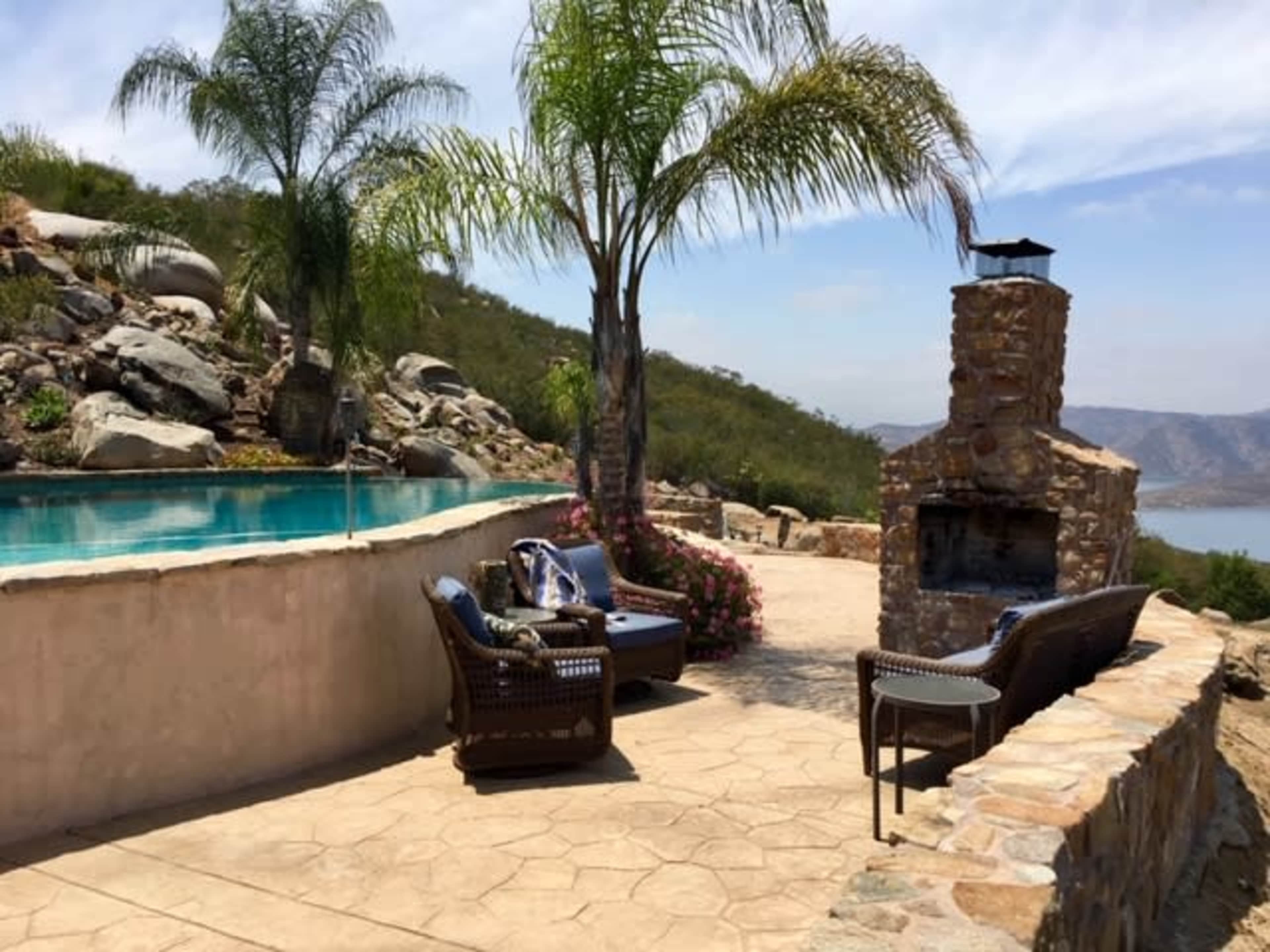 A stone patio with a fireplace, outdoor seating, and a swimming pool overlooking a mountainous landscape.