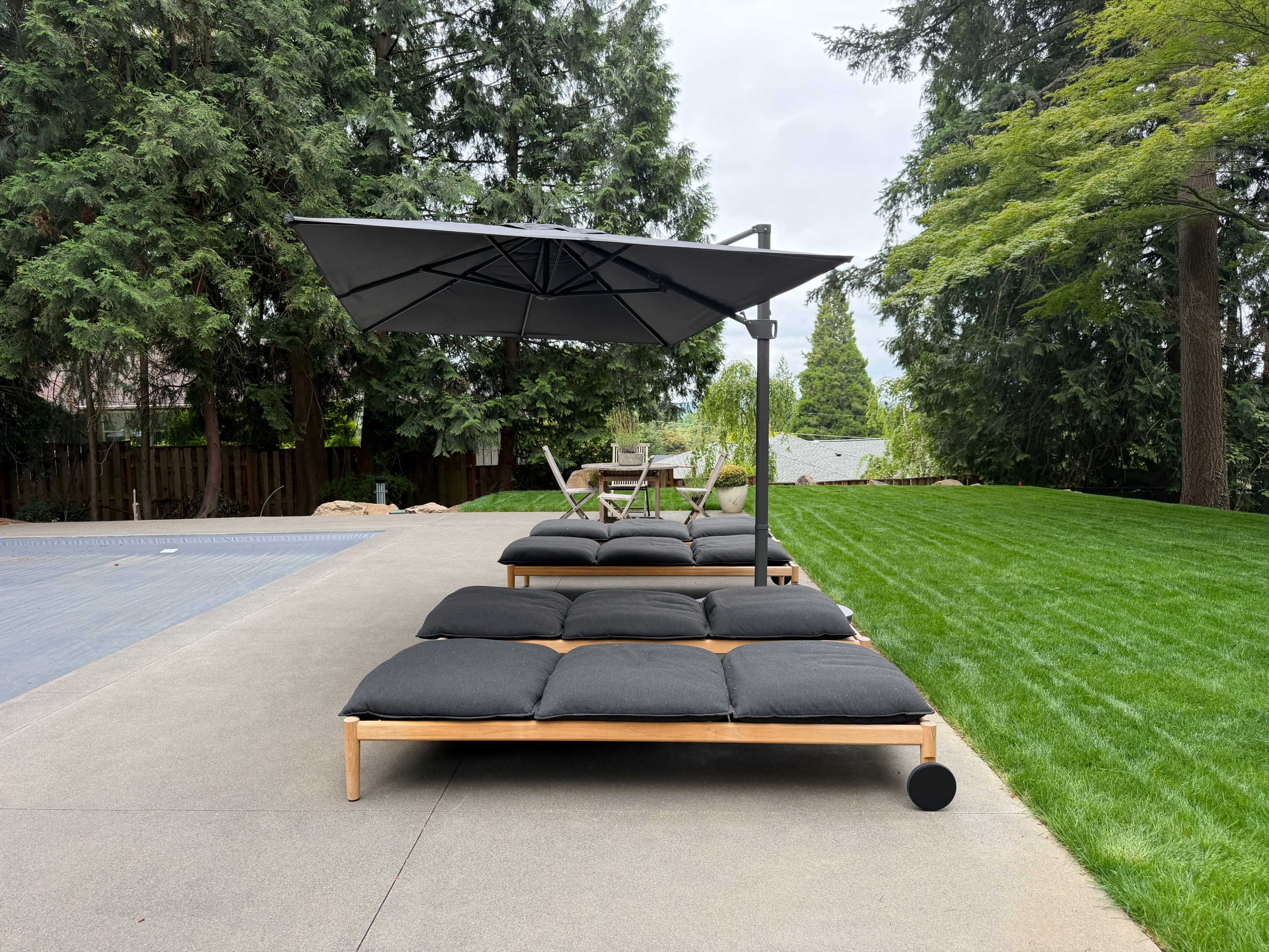 Two lounge beds with dark cushions and a large umbrella are positioned on a concrete patio, overlooking a grassy lawn.