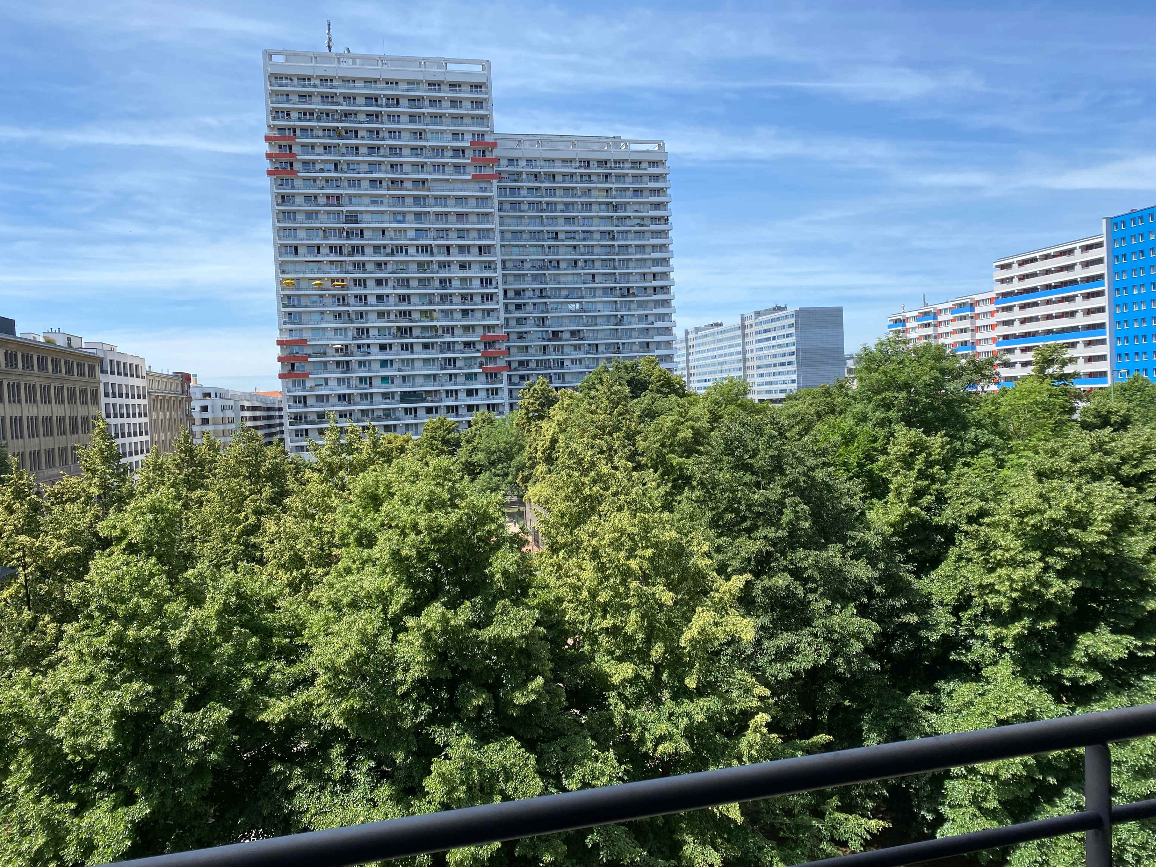 The image shows a view of tall residential buildings surrounded by lush green trees under a clear blue sky.