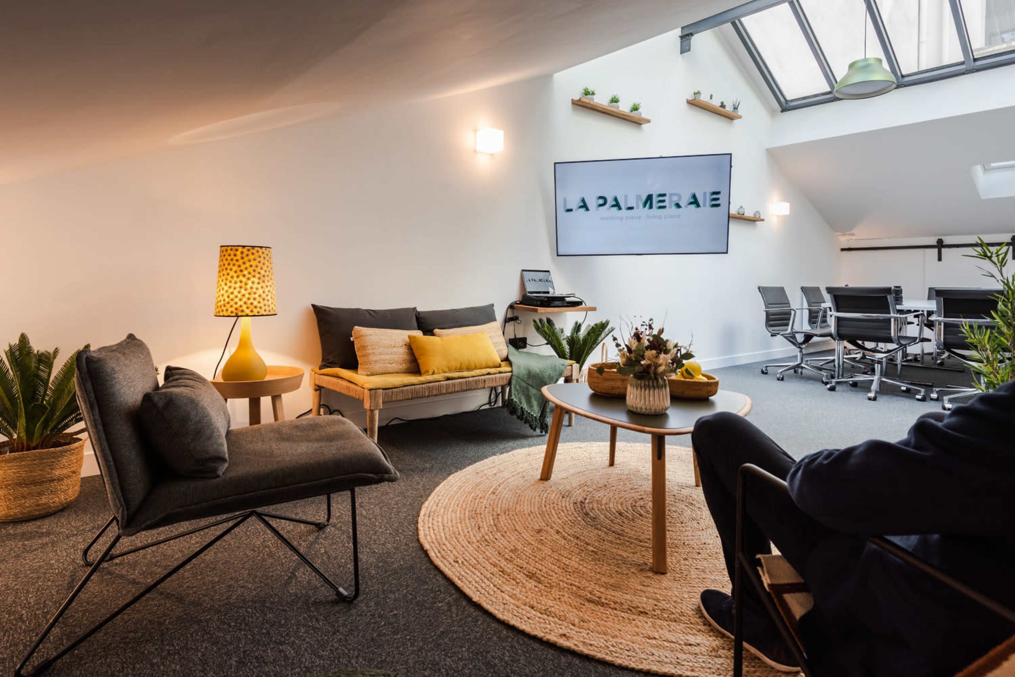 The image shows a modern office space with a cozy seating area featuring a light-colored sofa, a coffee table, and a large screen displaying "LA PALMERAIE," along with several plants and a conference table in the background.