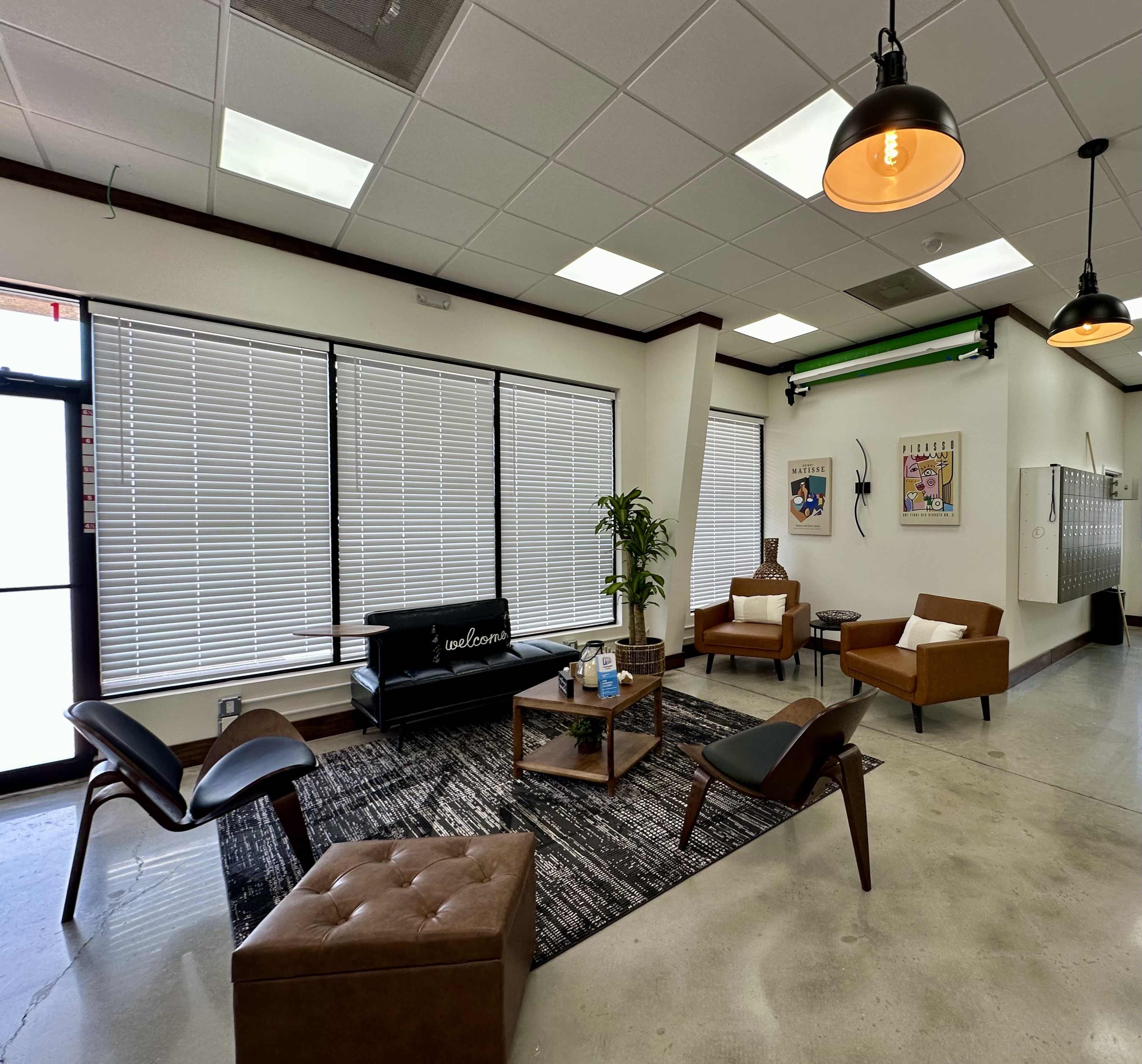 The image shows a modern lounge area with several seating options, large windows covered by blinds, and a coffee table on a patterned rug.