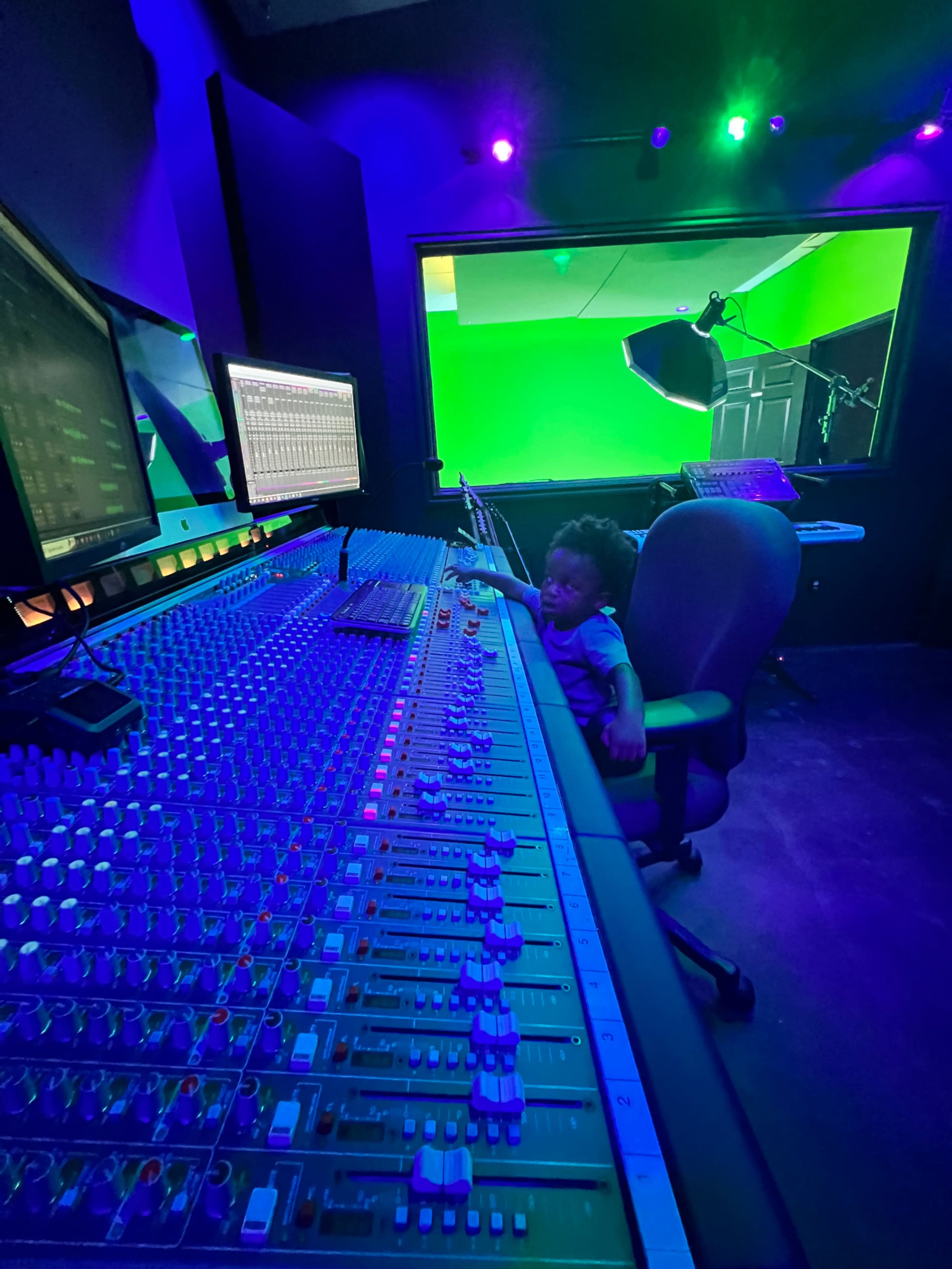 A child sits at a large sound mixing console in a dimly lit recording studio.