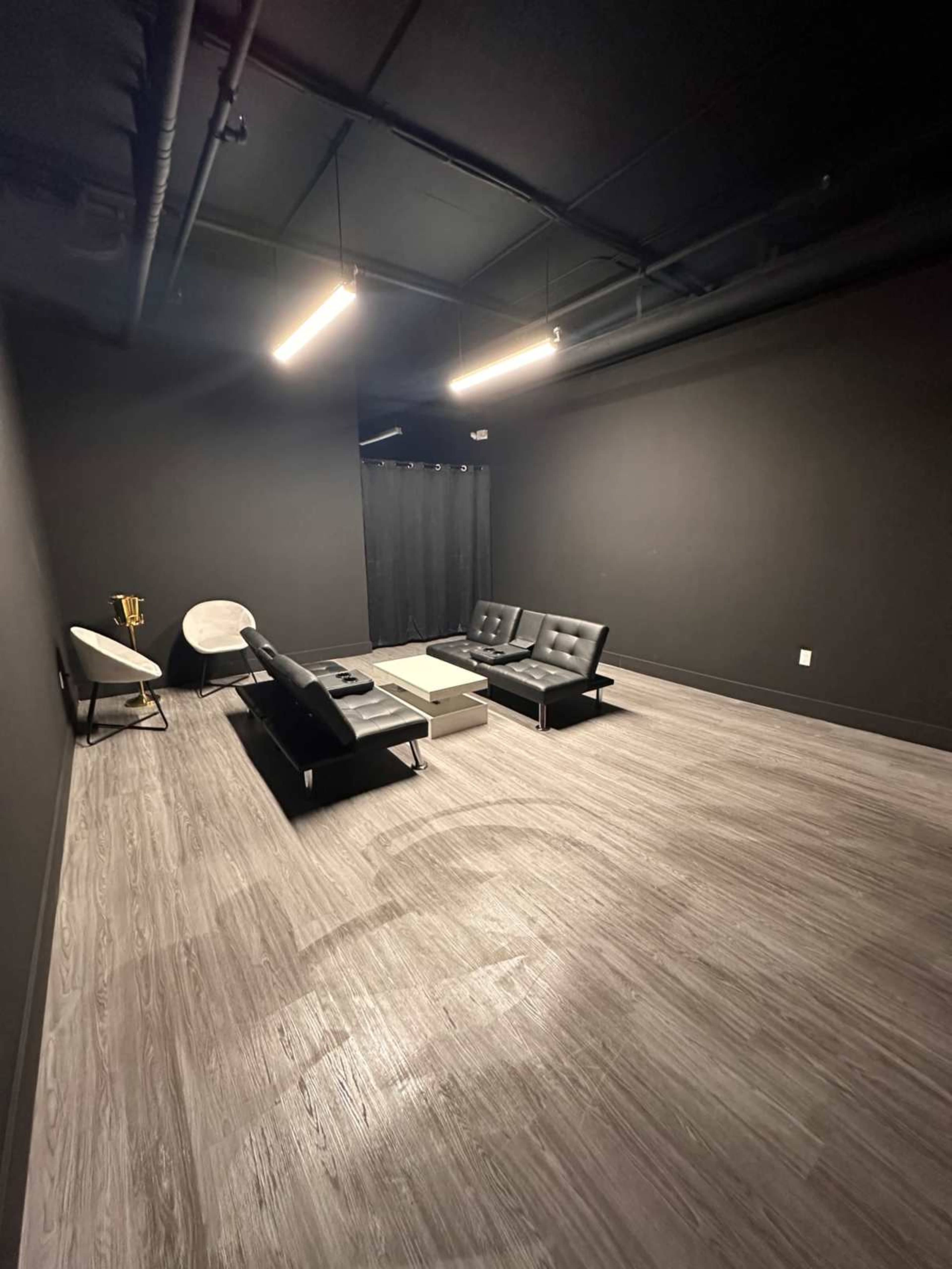 The image shows a spacious, dark room with black walls, featuring a black sectional sofa, a coffee table, and a white round chair.