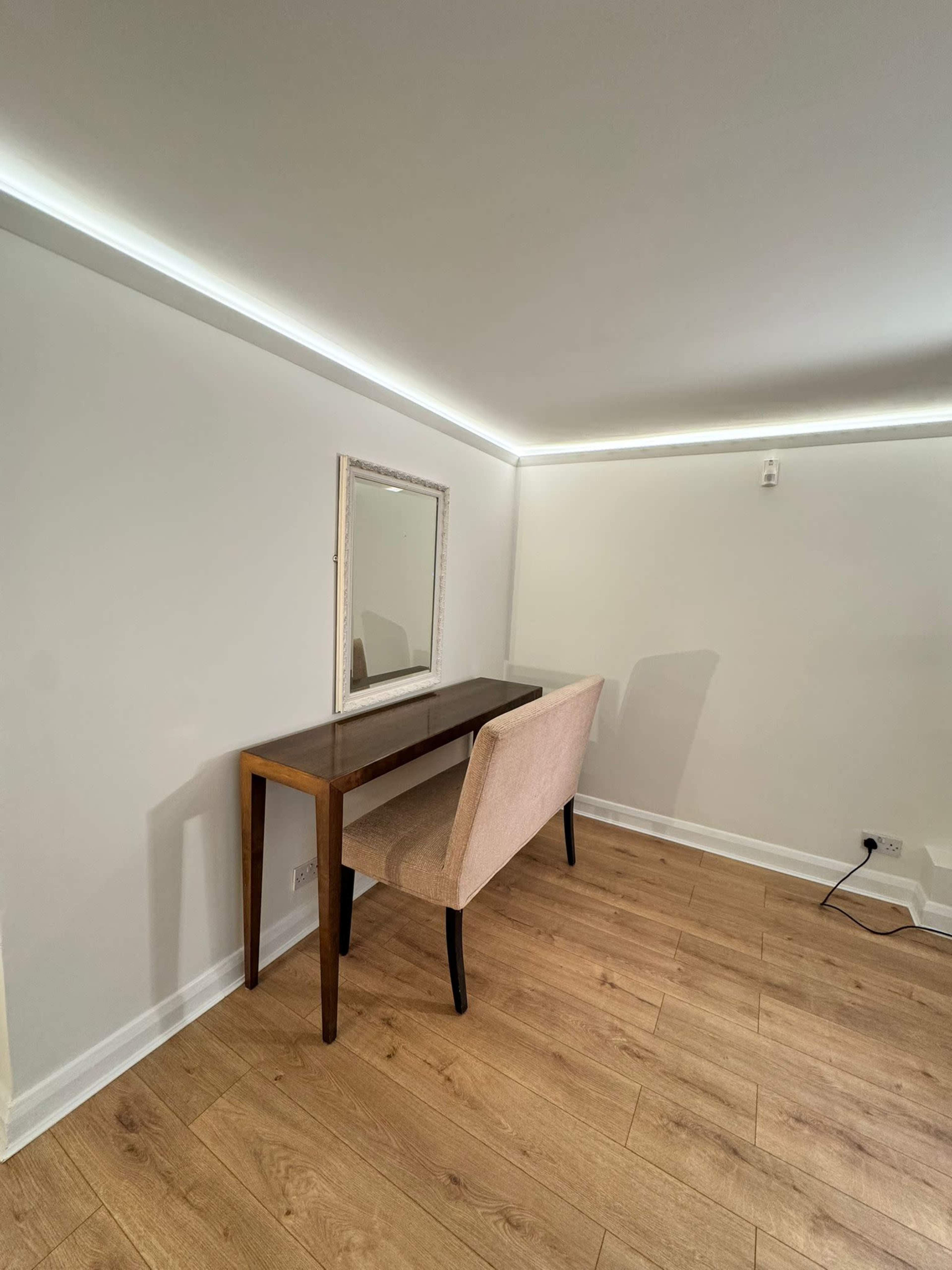 The image shows a small room featuring a wooden desk, a beige chair, and a mirror on the wall, illuminated by soft lighting along the ceiling.