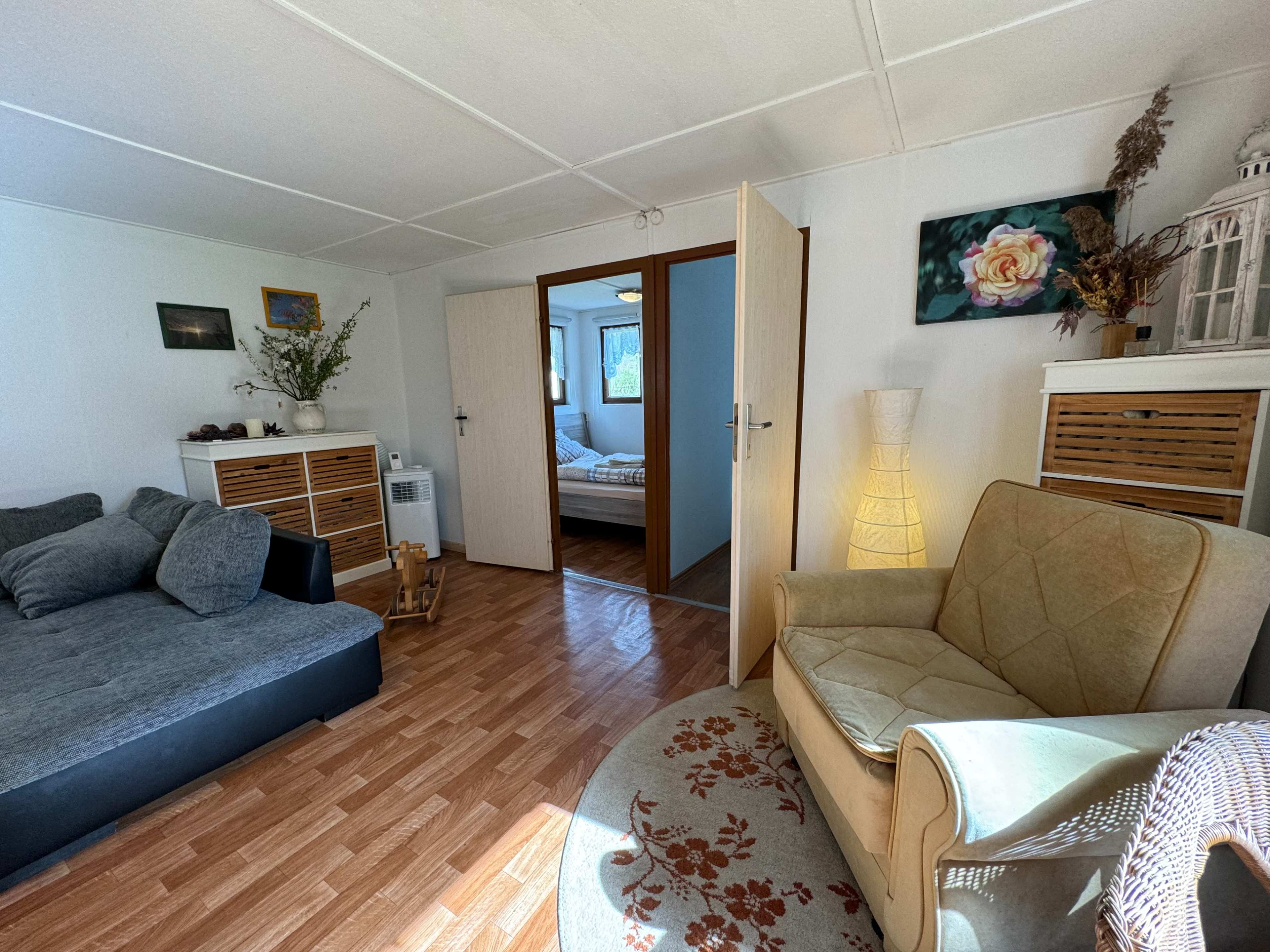 The image shows a cozy living room with a grey sectional sofa, a light-colored armchair, a decorative rug, and an open doorway leading to a bedroom.
