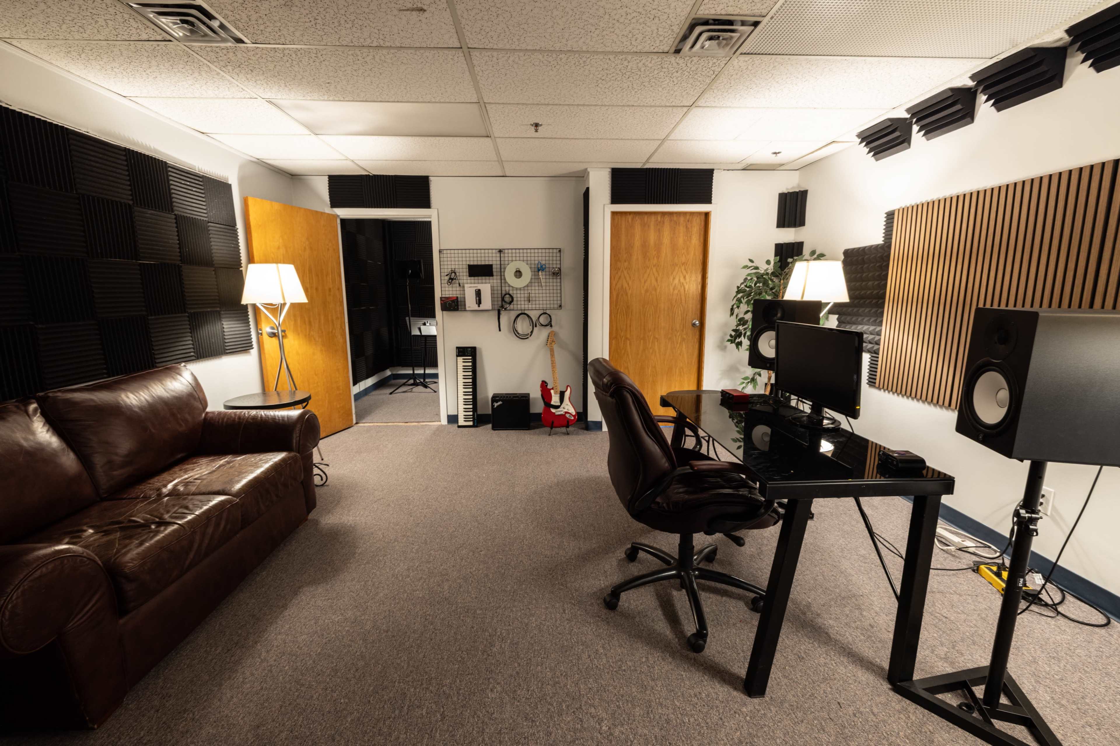 The image shows a soundproof studio room with a brown leather couch, a black desk with a monitor, and musical equipment on the walls, along with two doors leading to other rooms.