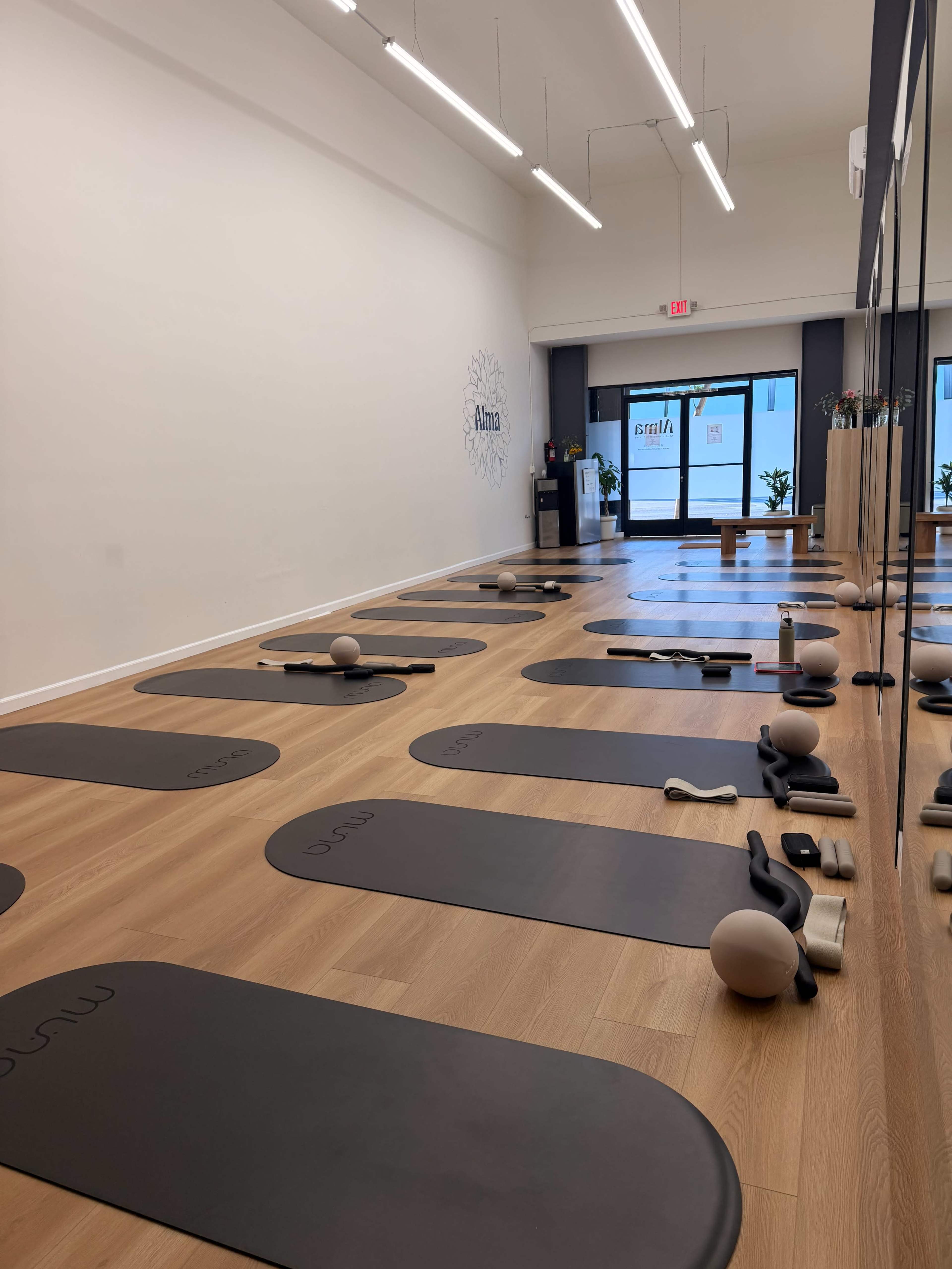 The image shows a clean and organized exercise studio with gray mats arranged neatly on a wooden floor, equipped with exercise props.