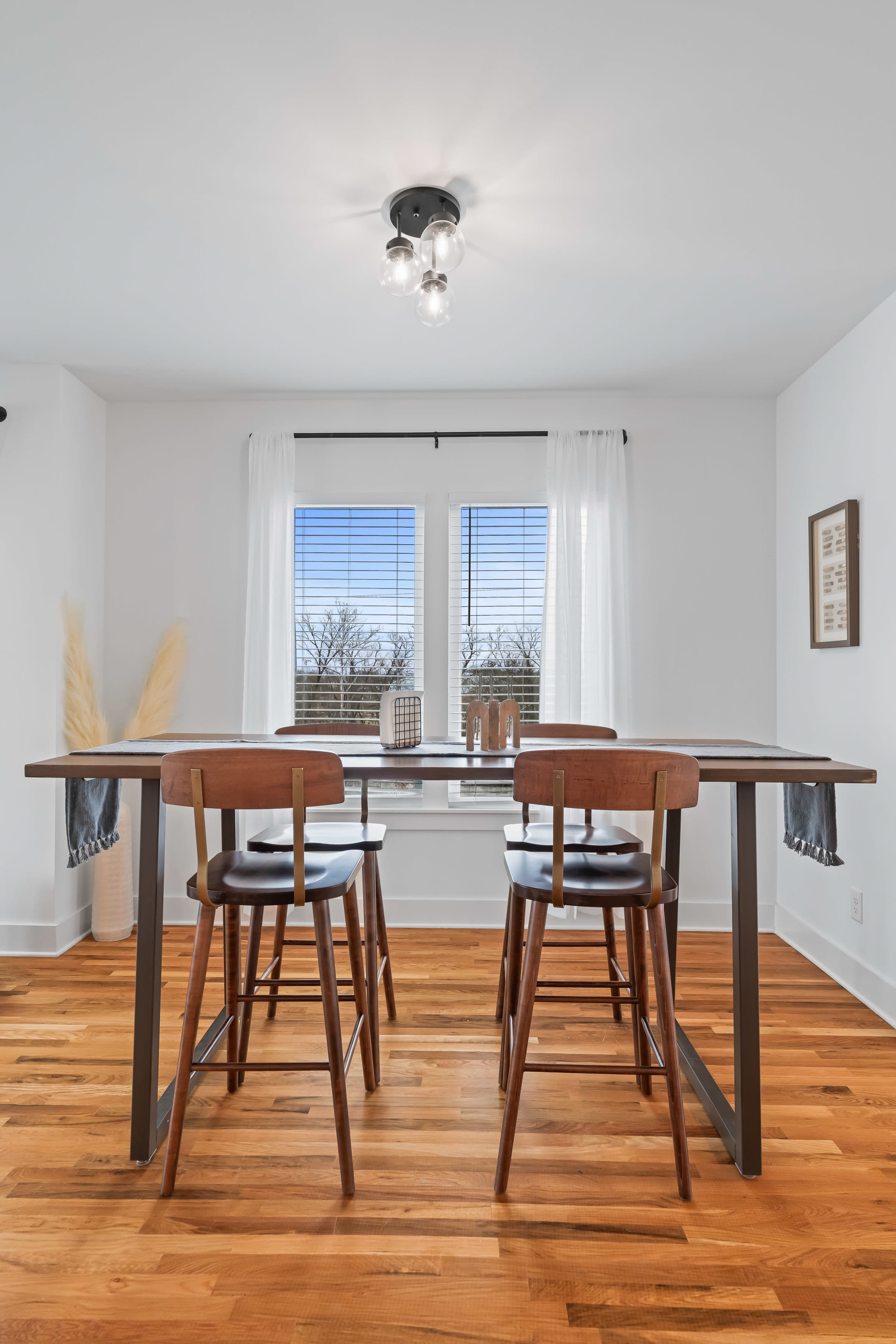 A modern dining area features a wooden table with two high-backed chairs, large windows revealing an outdoor view, and a light fixture overhead.