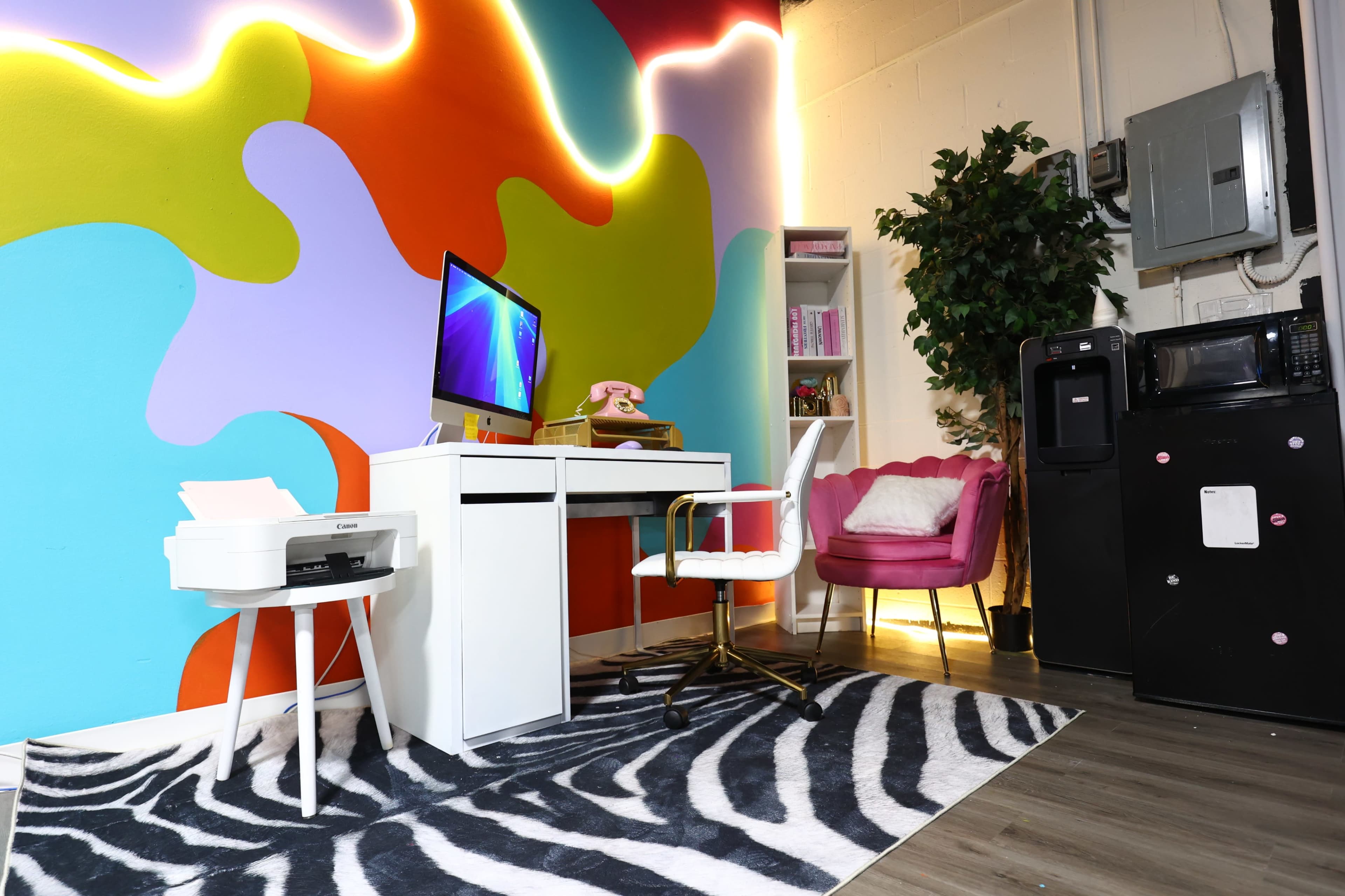 A brightly colored workspace features a desk with an iMac, a pink chair, and a plant beside a bookshelf, set against an abstract mural.