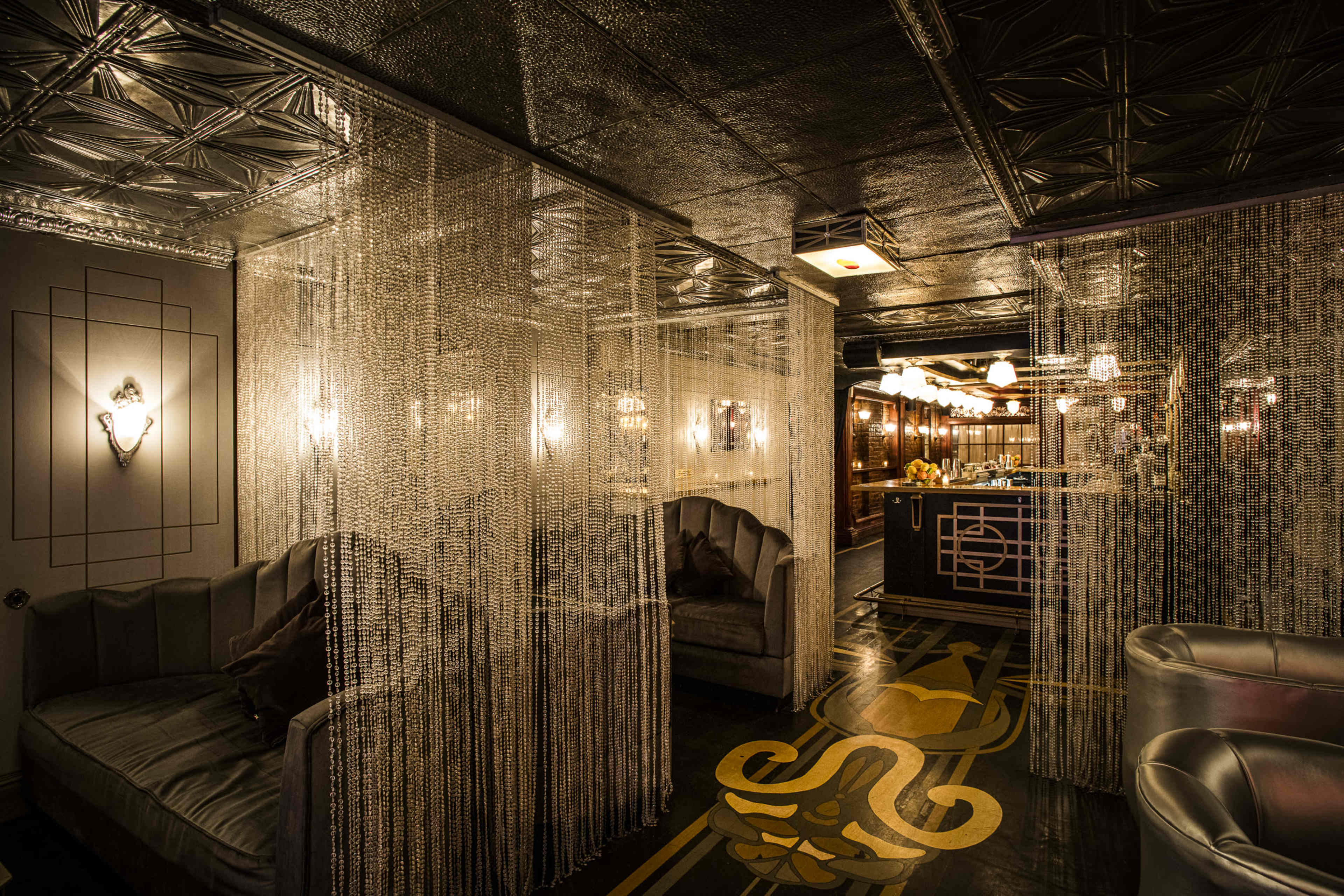 The image shows a dimly lit lounge with plush seating areas separated by decorative chain curtains, featuring a stylish bar in the background.