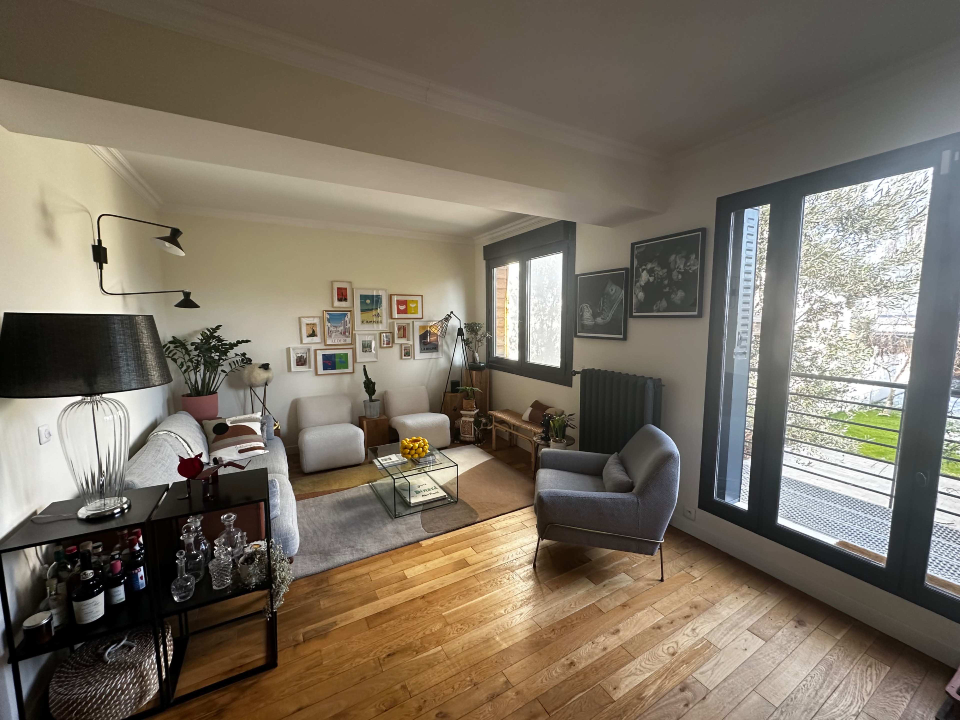The image shows a modern living room with wooden flooring, a grey sofa, a glass coffee table, and large windows allowing natural light to enter.