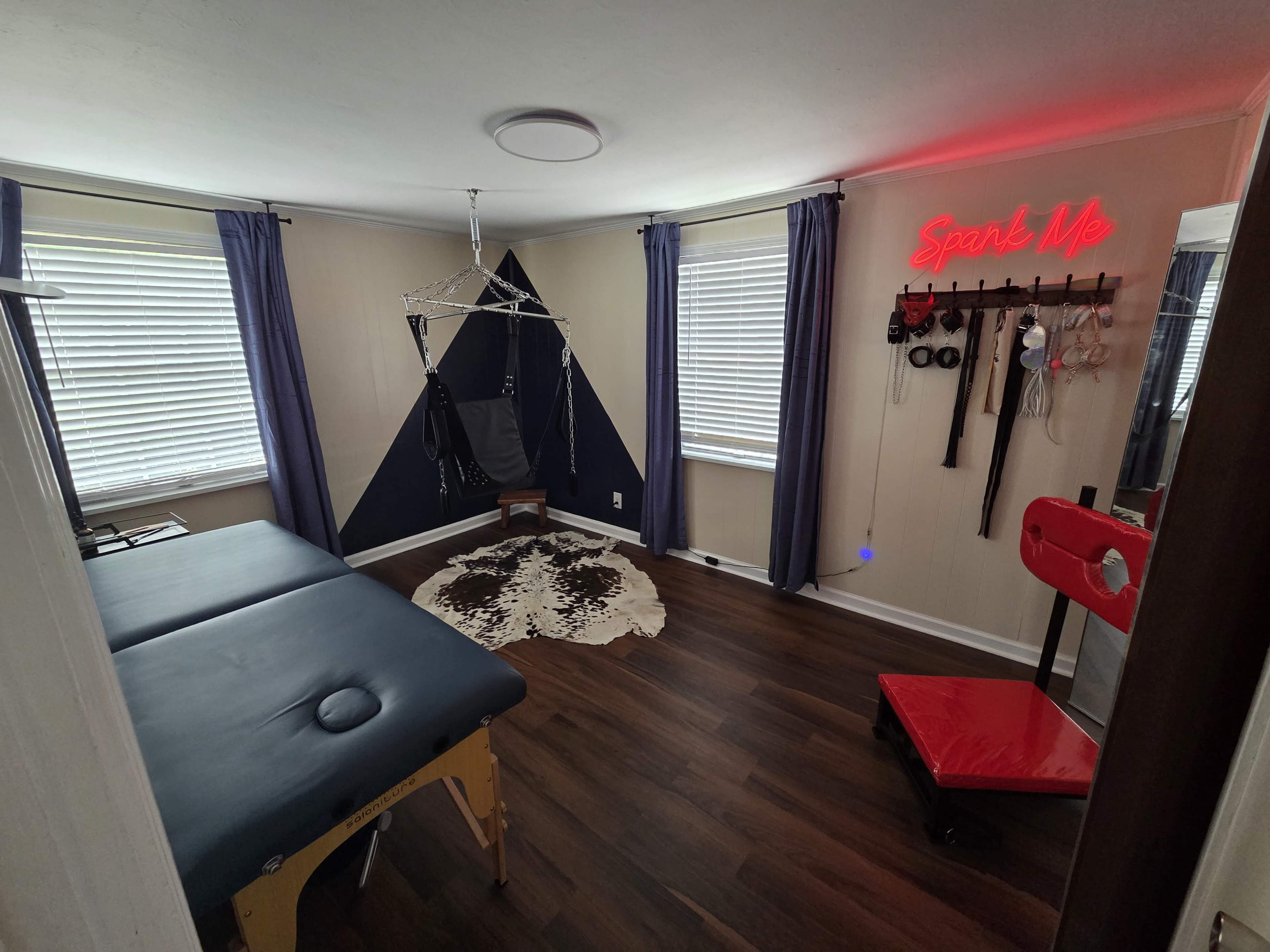 The room features a massage table, a black and white decorative wall, and various equipment on display, including a red chair and a neon sign.