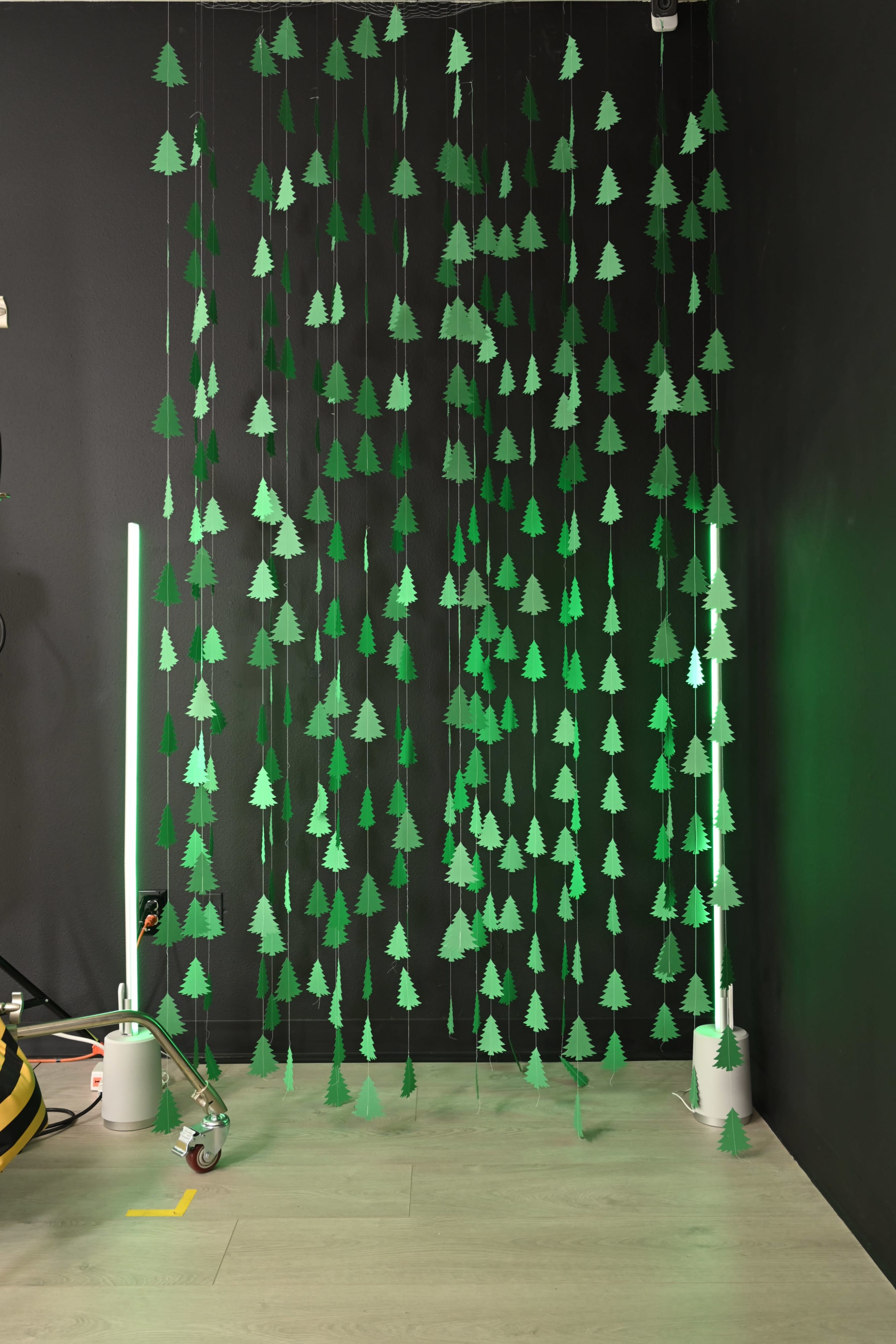 The image features a wall covered with hanging green cutouts of trees, with two vertical light fixtures on the floor beside them.