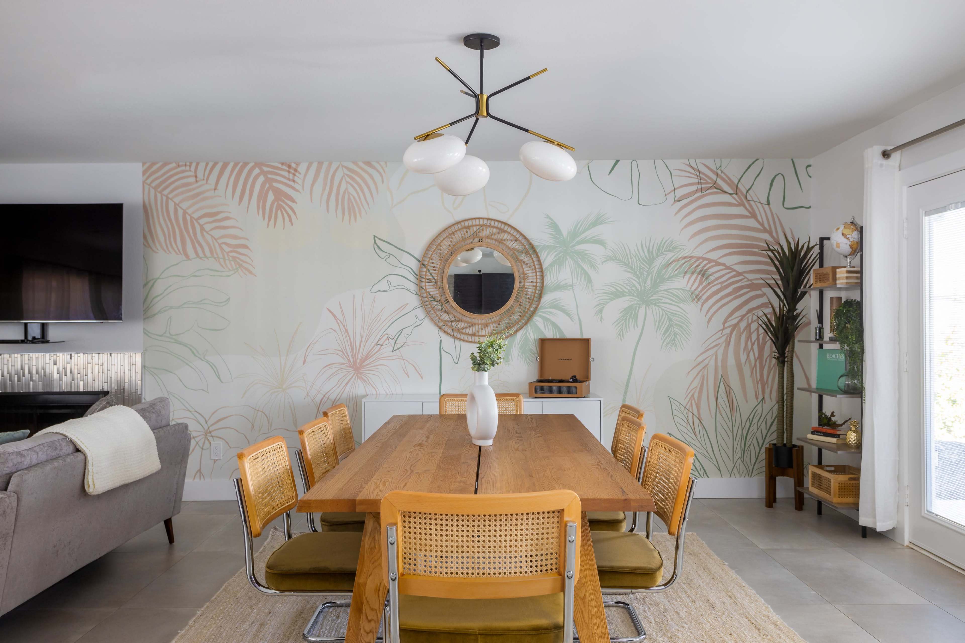 A wooden dining table with yellow chairs is centered in a modern dining area featuring a palm-themed mural and a circular mirror on the wall.