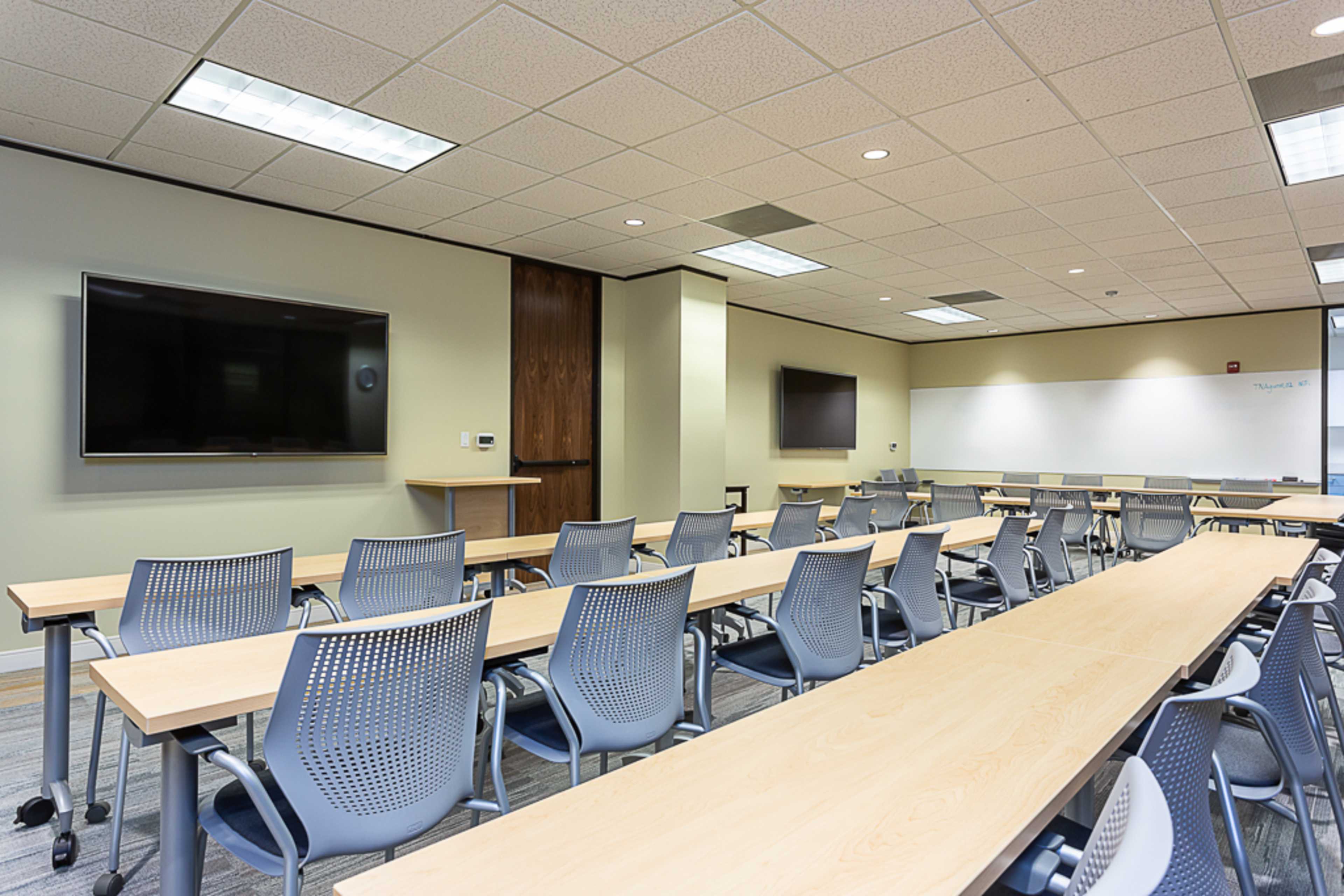 A spacious classroom setup with rows of tables and chairs, large wall-mounted screens, and a whiteboard.