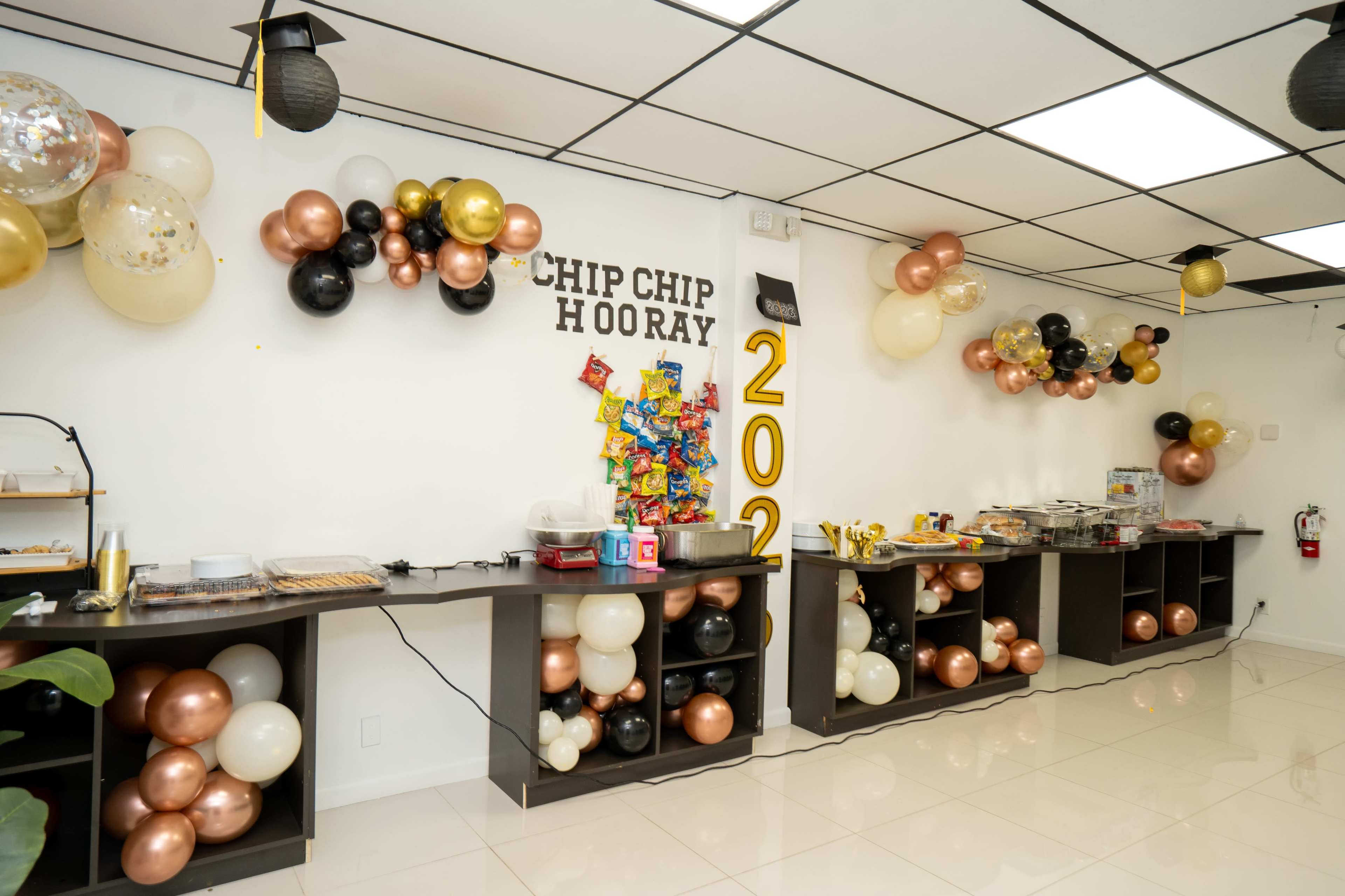 The image shows a festively decorated room featuring a "CHIP CHIP HOORAY" banner, colorful balloons, and a food display on black tables.