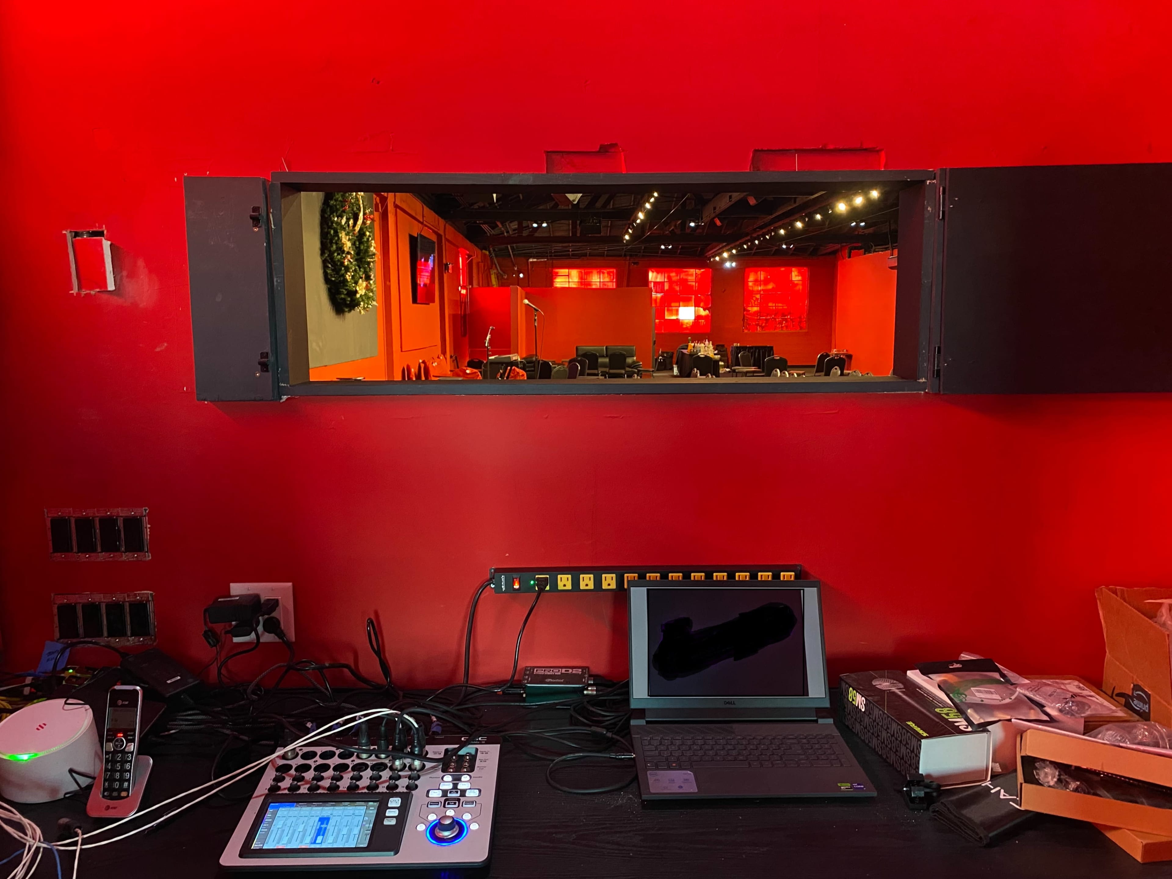 The image shows a control station with electronic equipment, including a mixer and a laptop, set against a vibrant red wall, overlooking a separate room with seating.