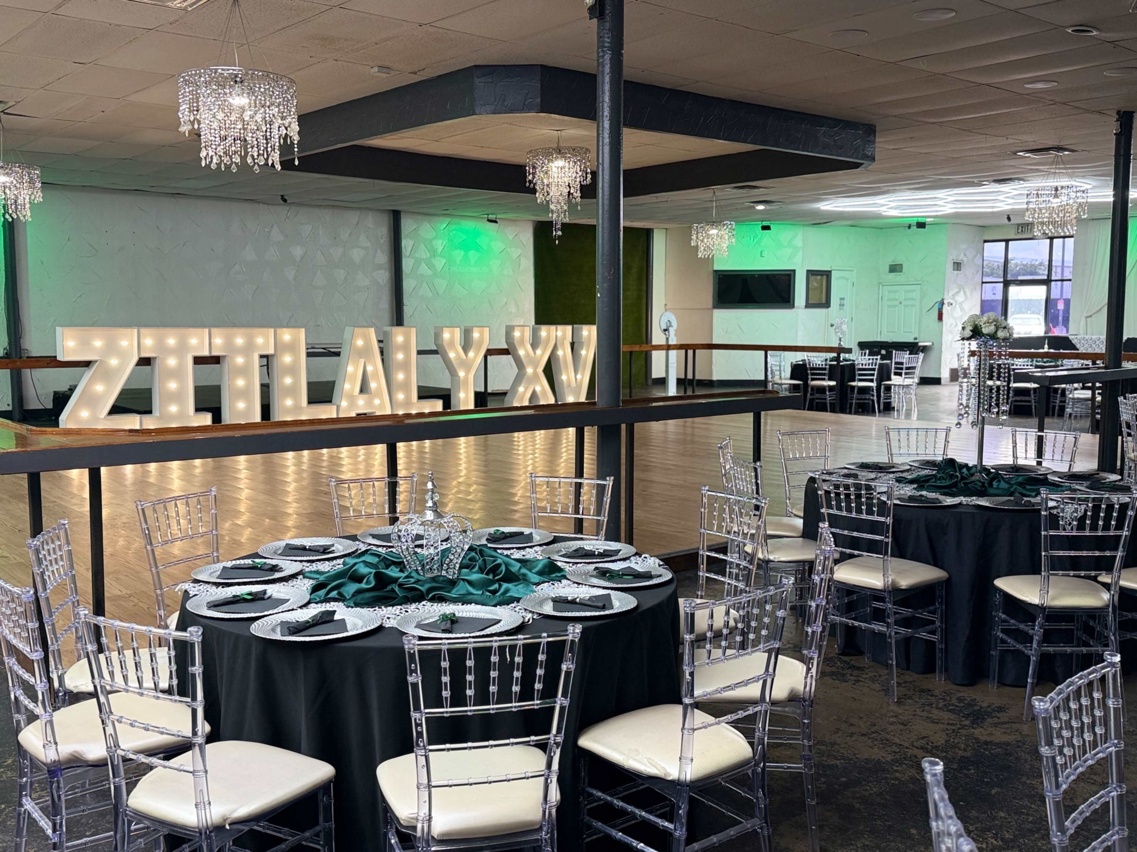 The image shows an elegantly decorated event space featuring round tables with black tablecloths, clear chiavari chairs, and a large illuminated sign that reads "ZITALY XV."