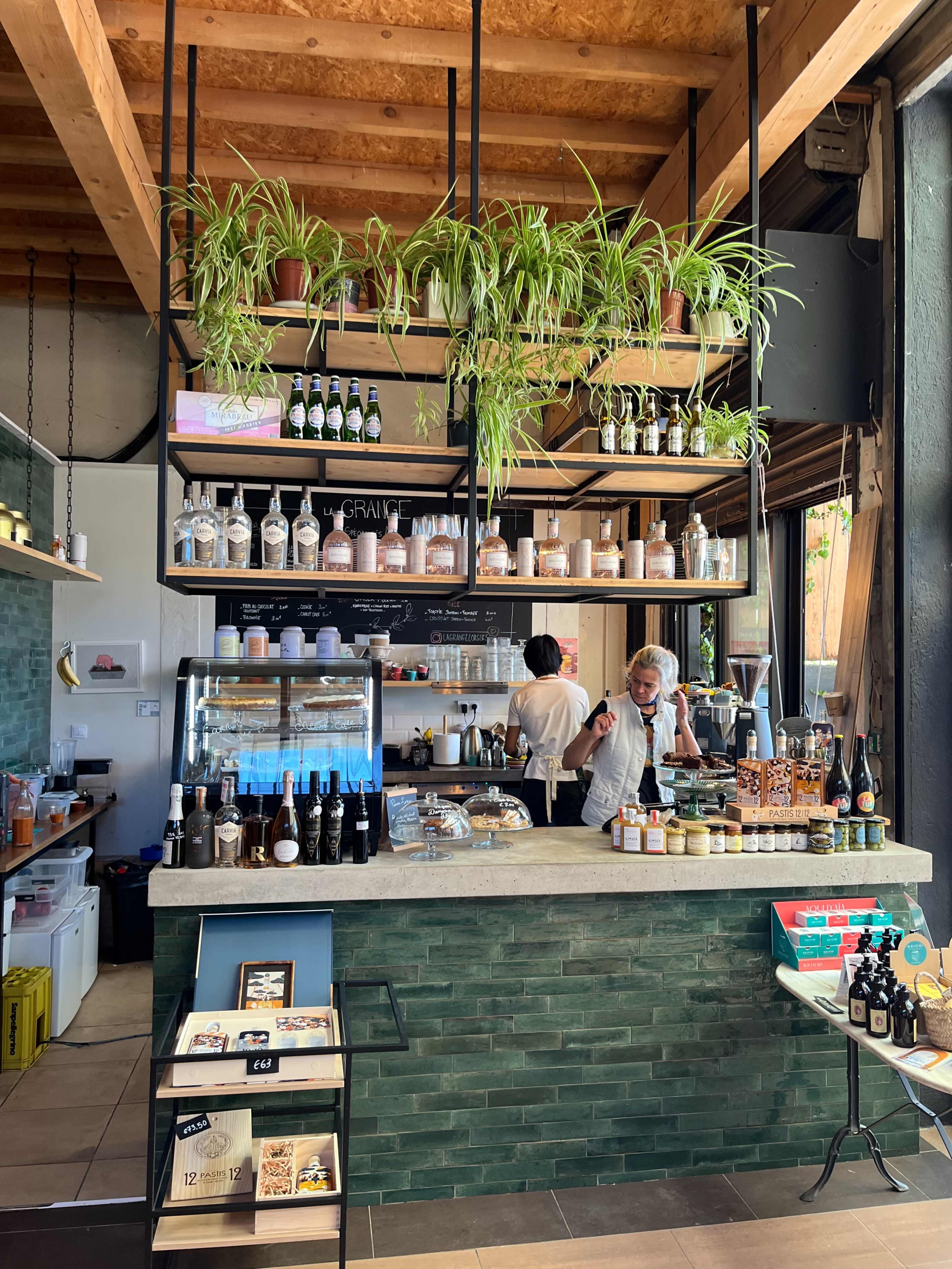 The image shows a modern cafe interior with a counter displaying various beverages and snacks, surrounded by green plants and a barista preparing drinks.