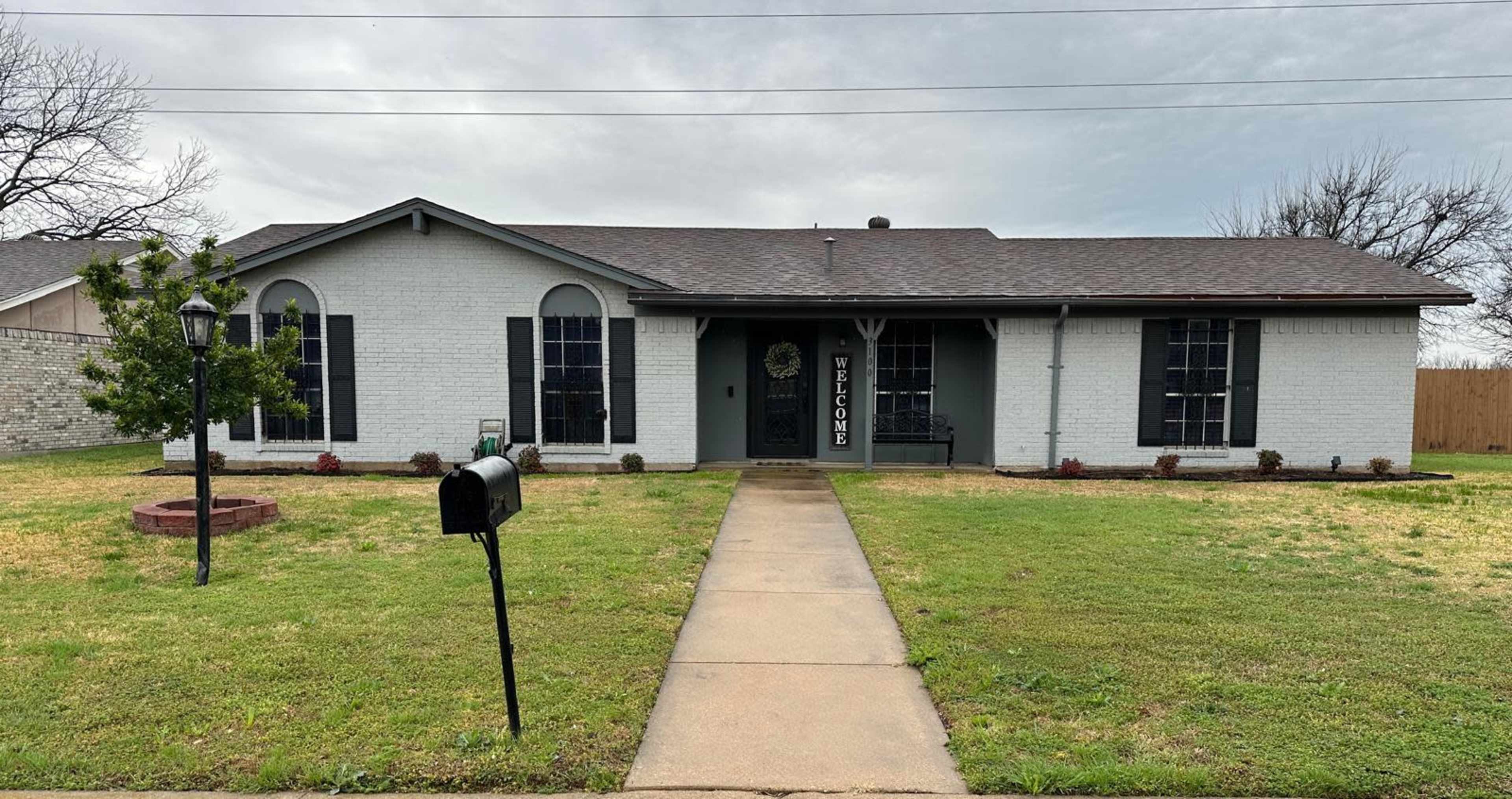 The image shows a single-story, white-brick house with dark shutters, a well-maintained front lawn, and a welcoming entryway with a sign.