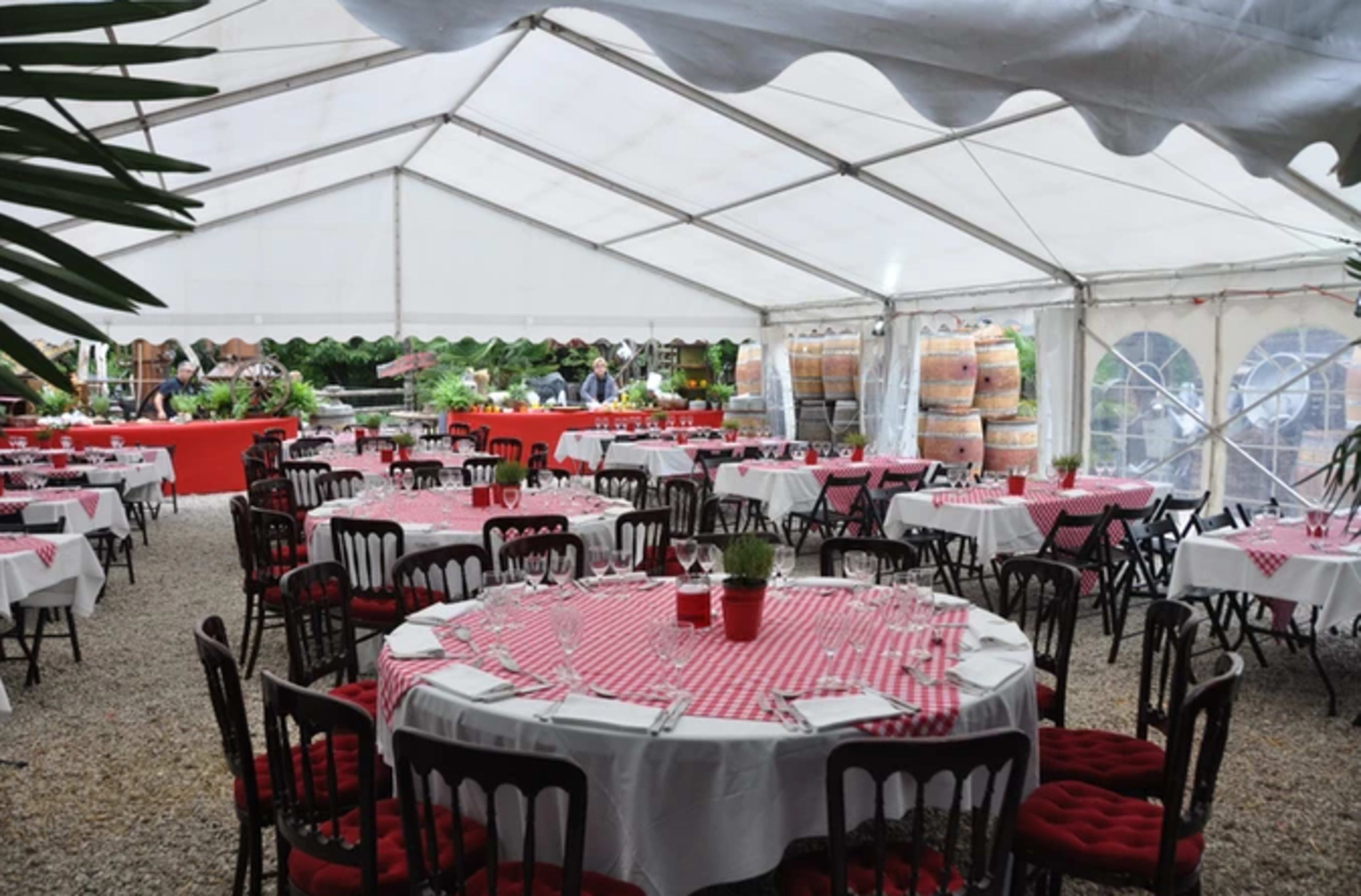 A large tented dining area features round tables dressed in red and white checkered cloths, surrounded by chairs, with food displays and wine barrels in the background.