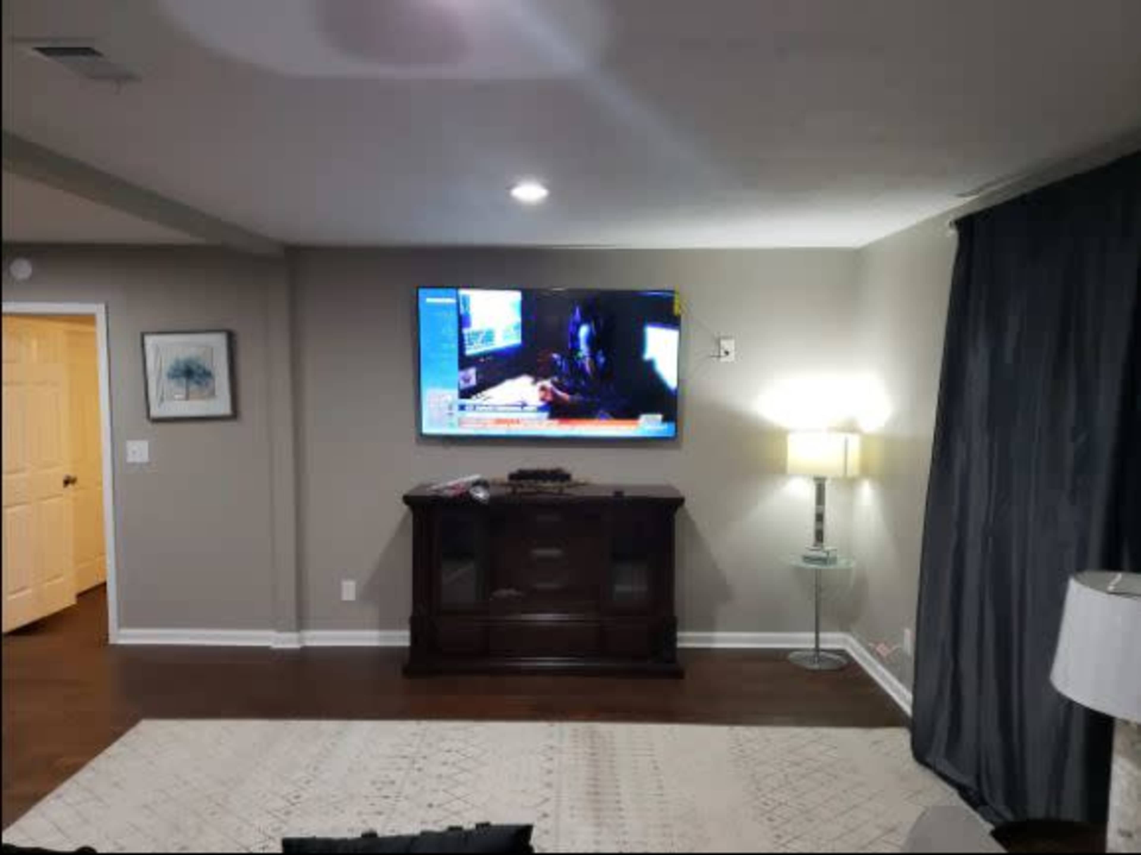 A living room with a wall-mounted TV displaying a news program, a dark curtain, a lamp beside a wooden cabinet, and a light-colored rug on the floor.
