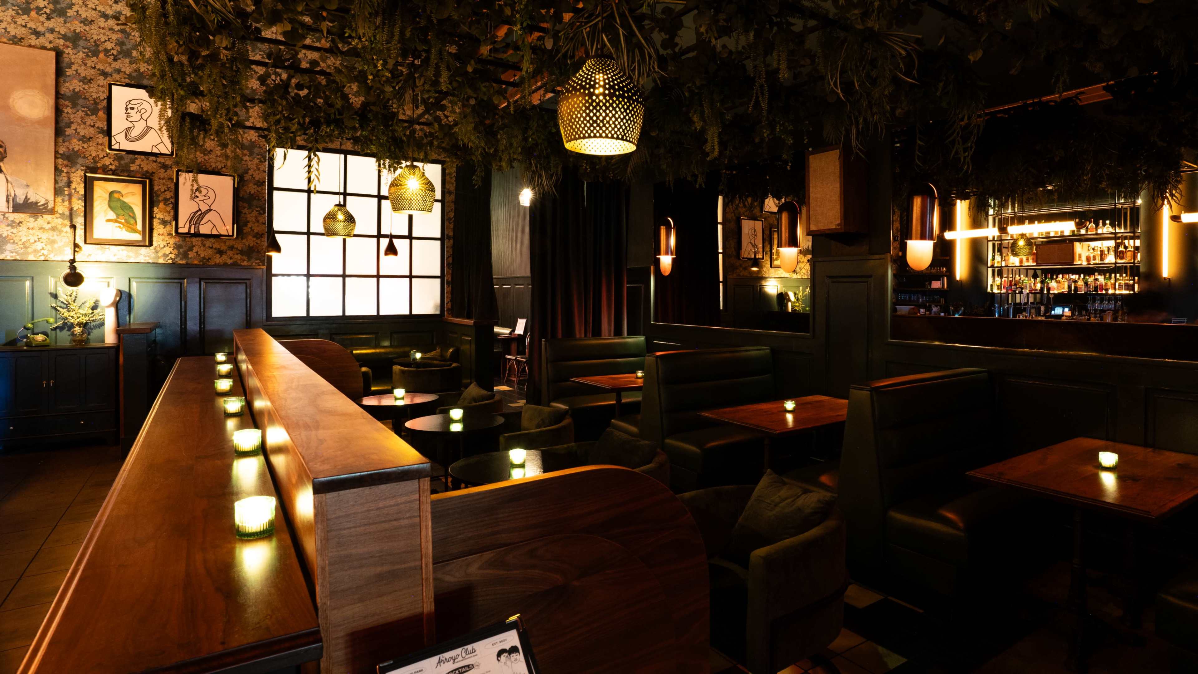 The image shows a dimly lit restaurant interior with wooden tables, green upholstered seating, and decorative wall art under a canopy of foliage.