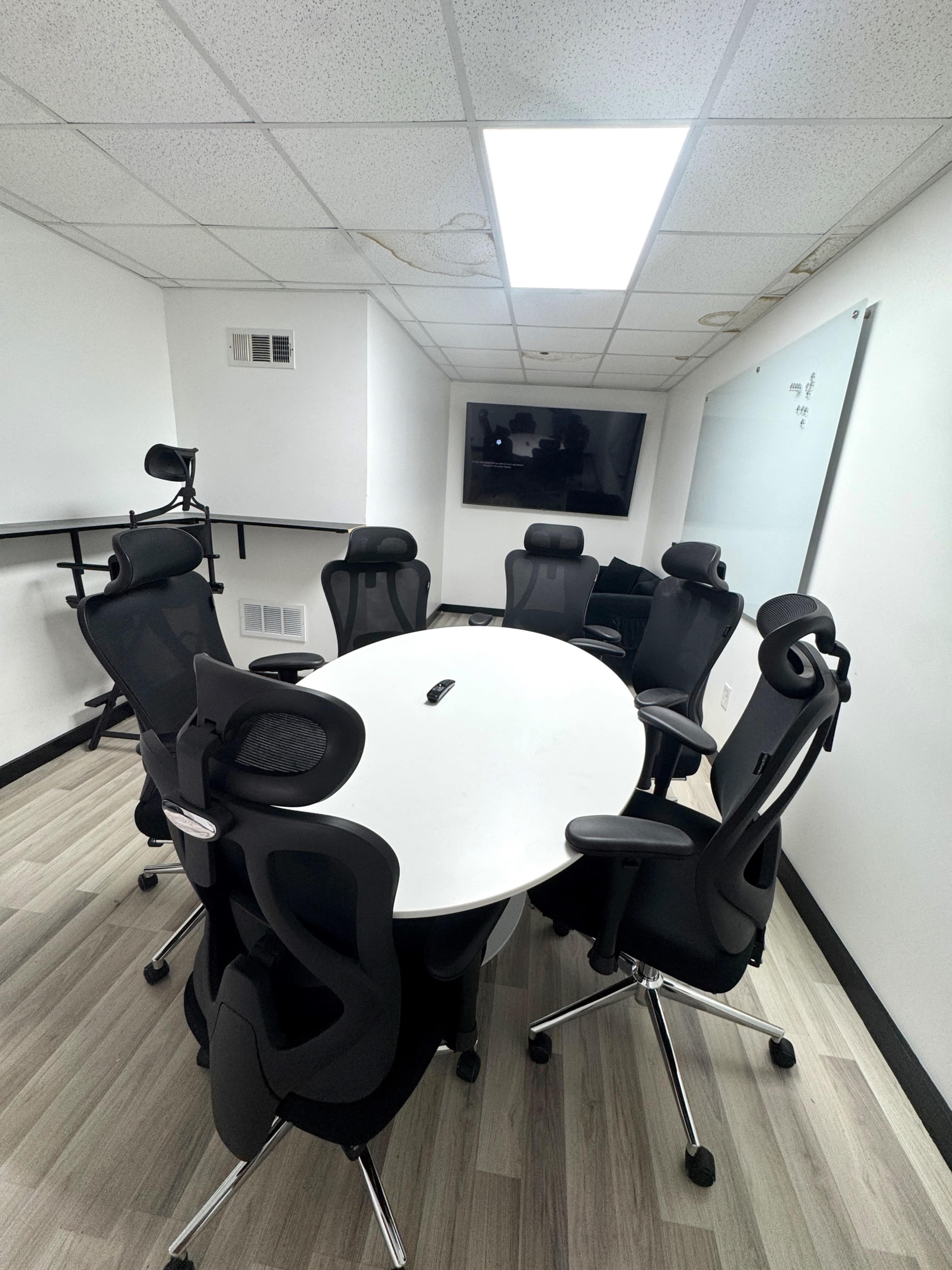 A conference room features a white oval table surrounded by six black ergonomic chairs, with a television screen mounted on the wall.
