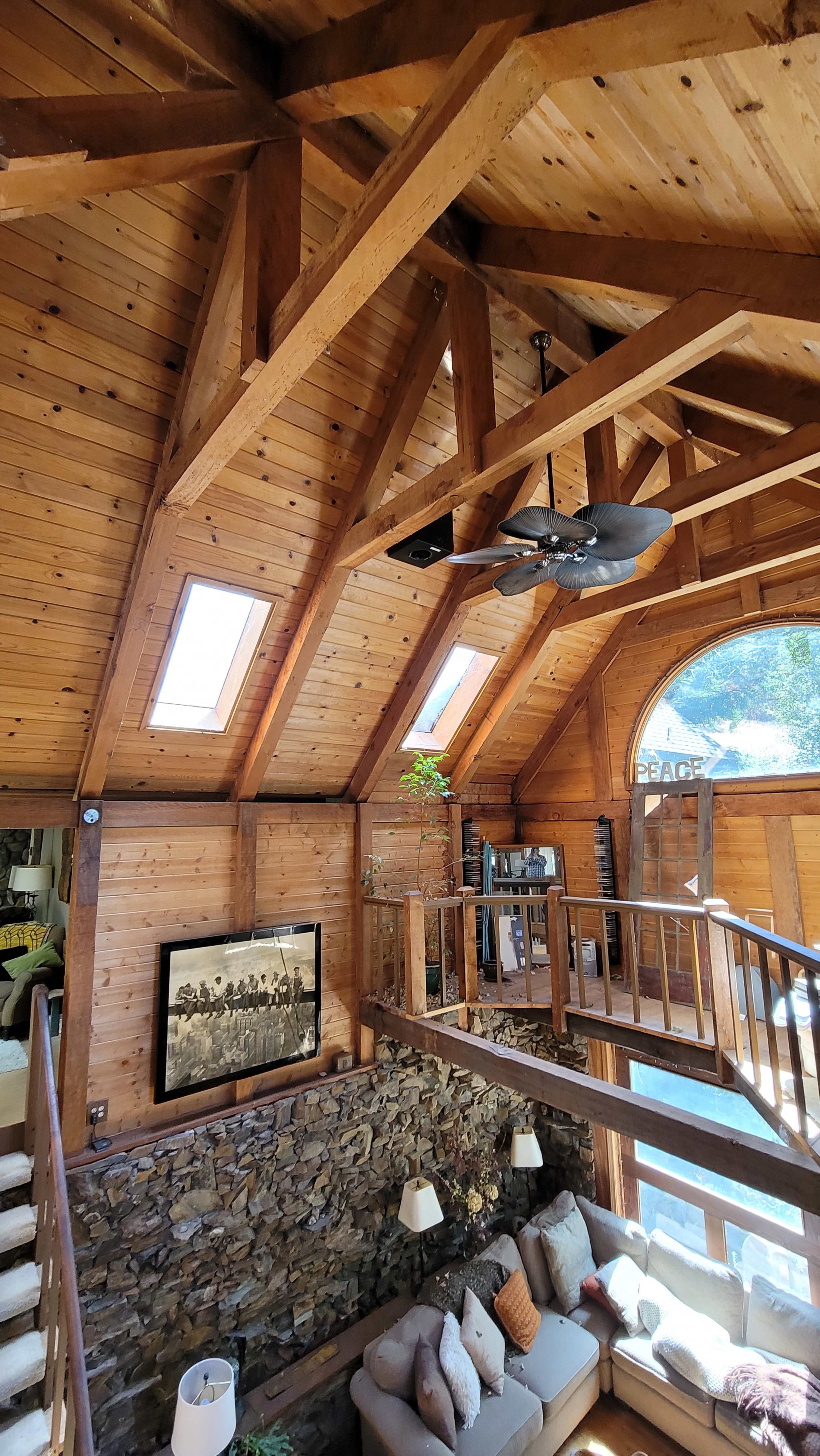 A wooden interior of a house featuring exposed beams, a stone wall, and a cozy seating area with large windows allowing natural light to enter.