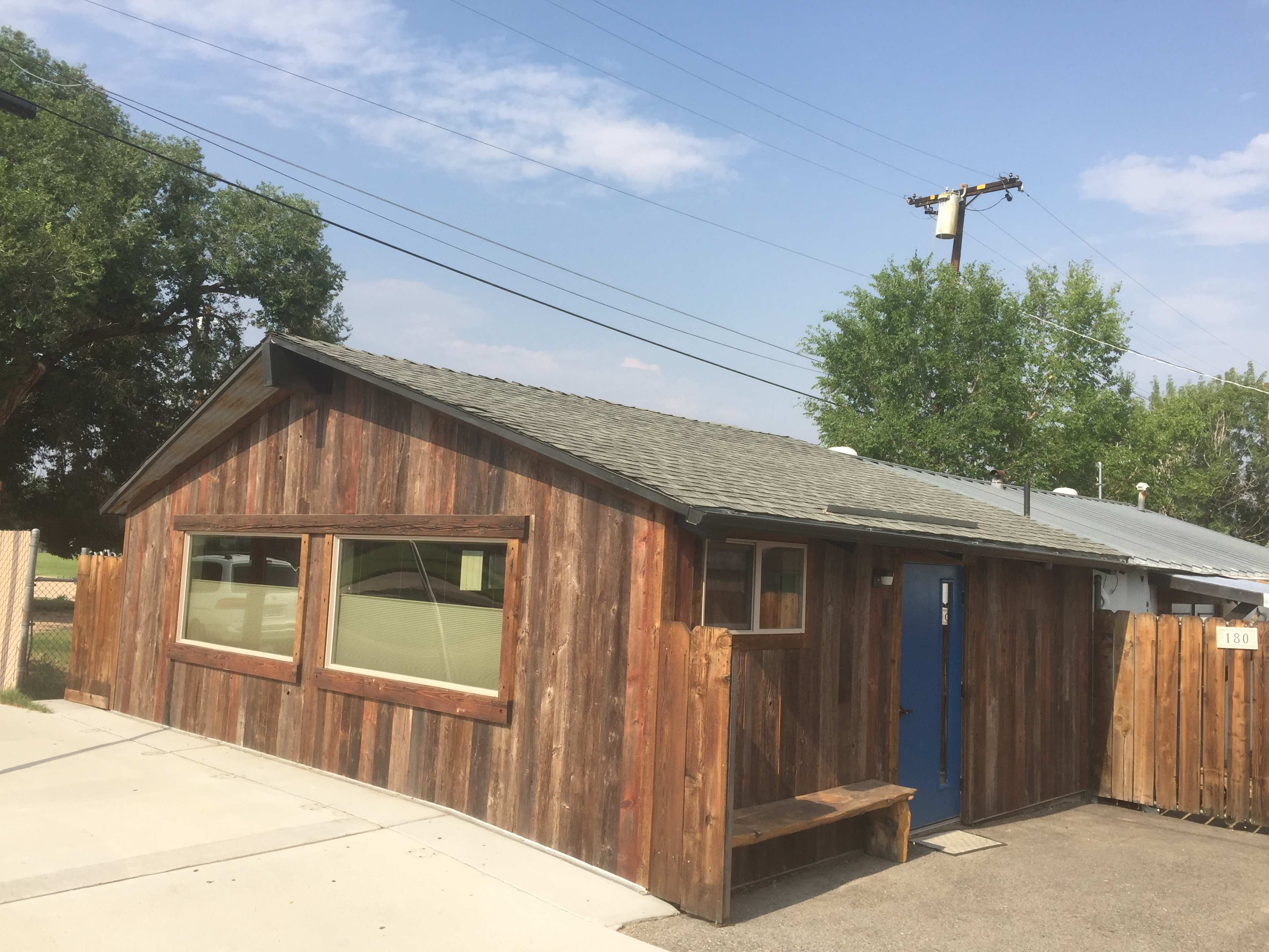 A wooden structure with a blue door and large windows stands in an open area, bordered by a wooden fence and surrounded by trees.