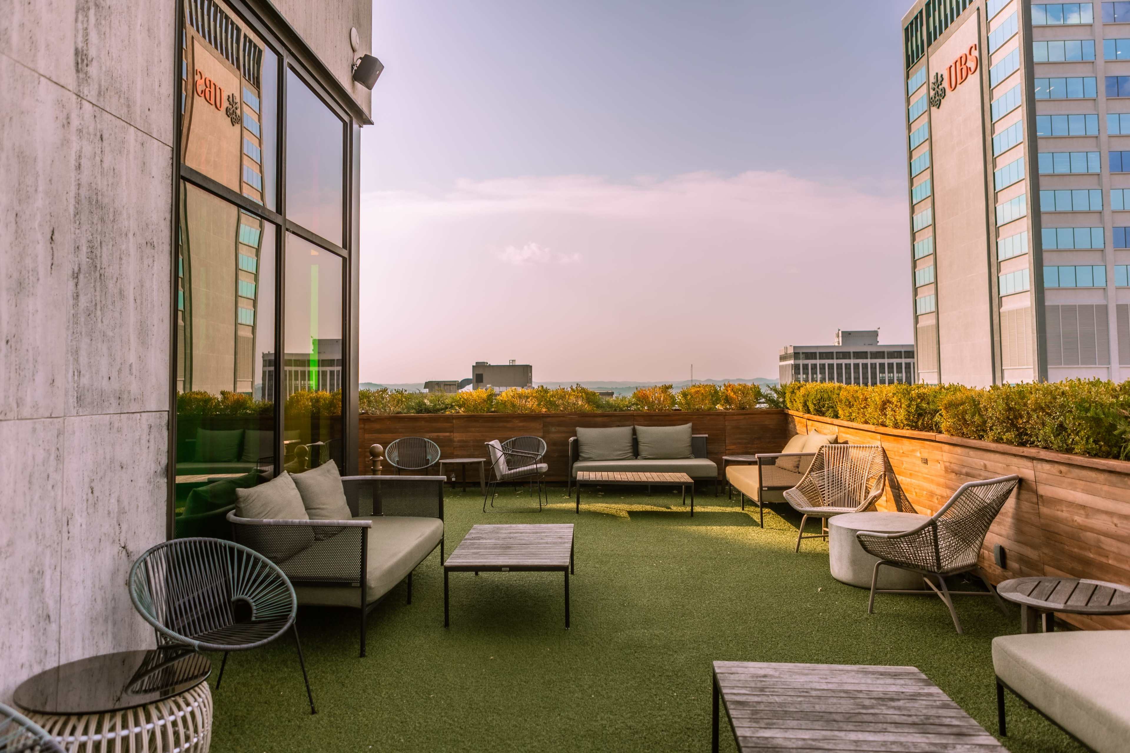 A rooftop lounge area with modern seating and greenery, set against a city skyline.