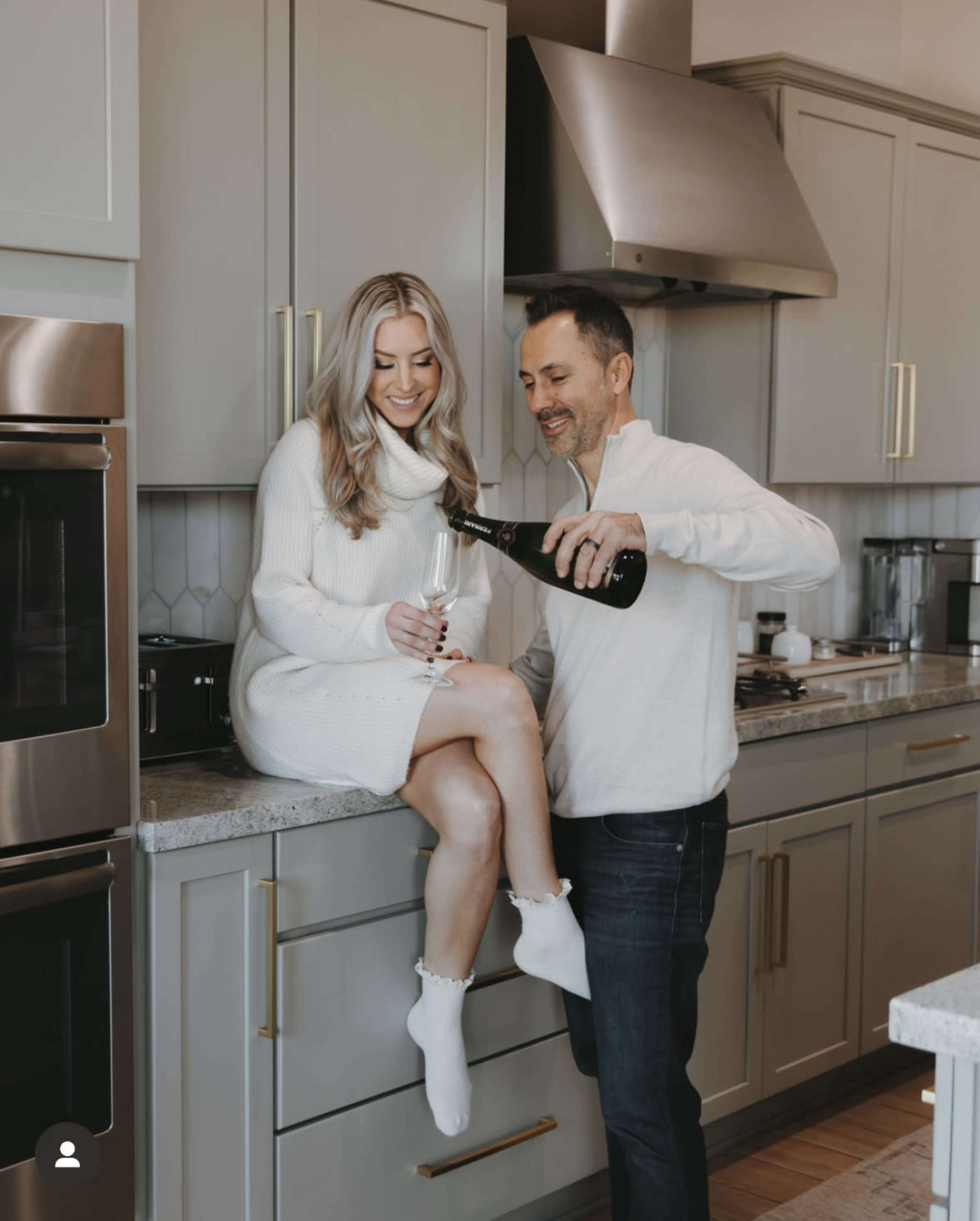 A man pours champagne into a glass held by a woman sitting on a kitchen countertop.