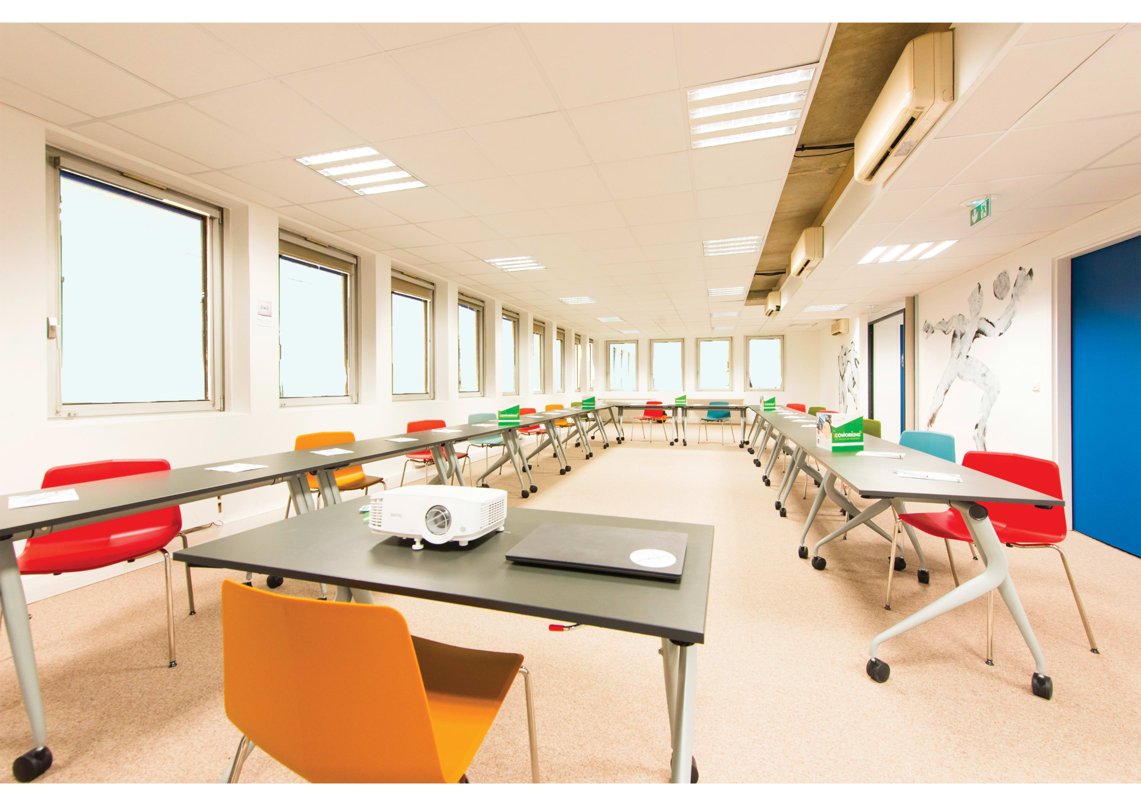 A bright, modern classroom features multiple tables arranged in a U-shape with colorful chairs and a projector in the center.
