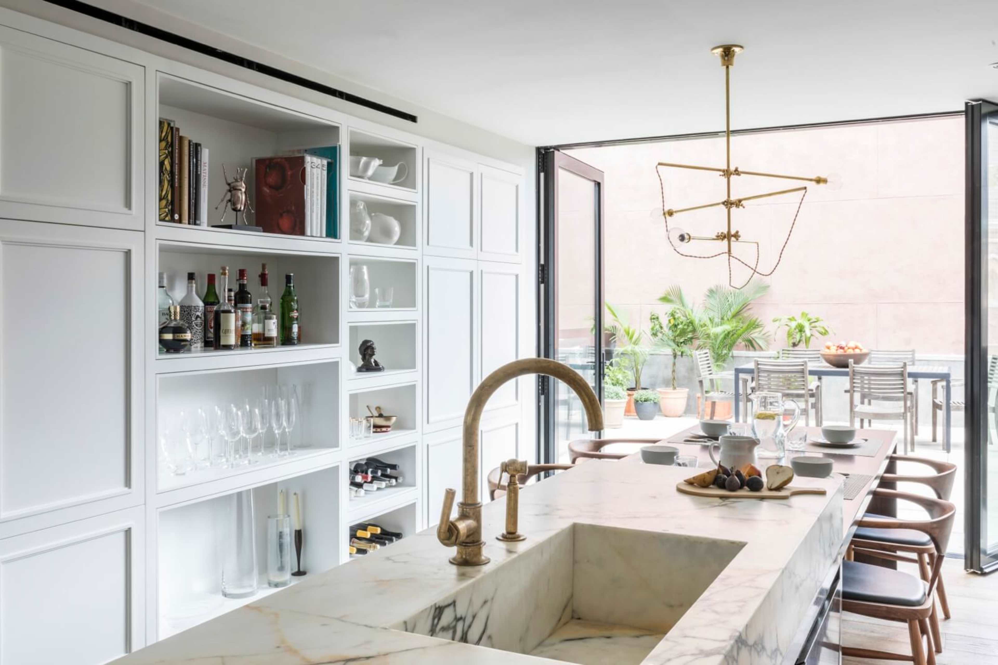A modern kitchen with a marble island, open shelving displaying glassware and bottles, and large sliding doors leading to an outdoor dining area.