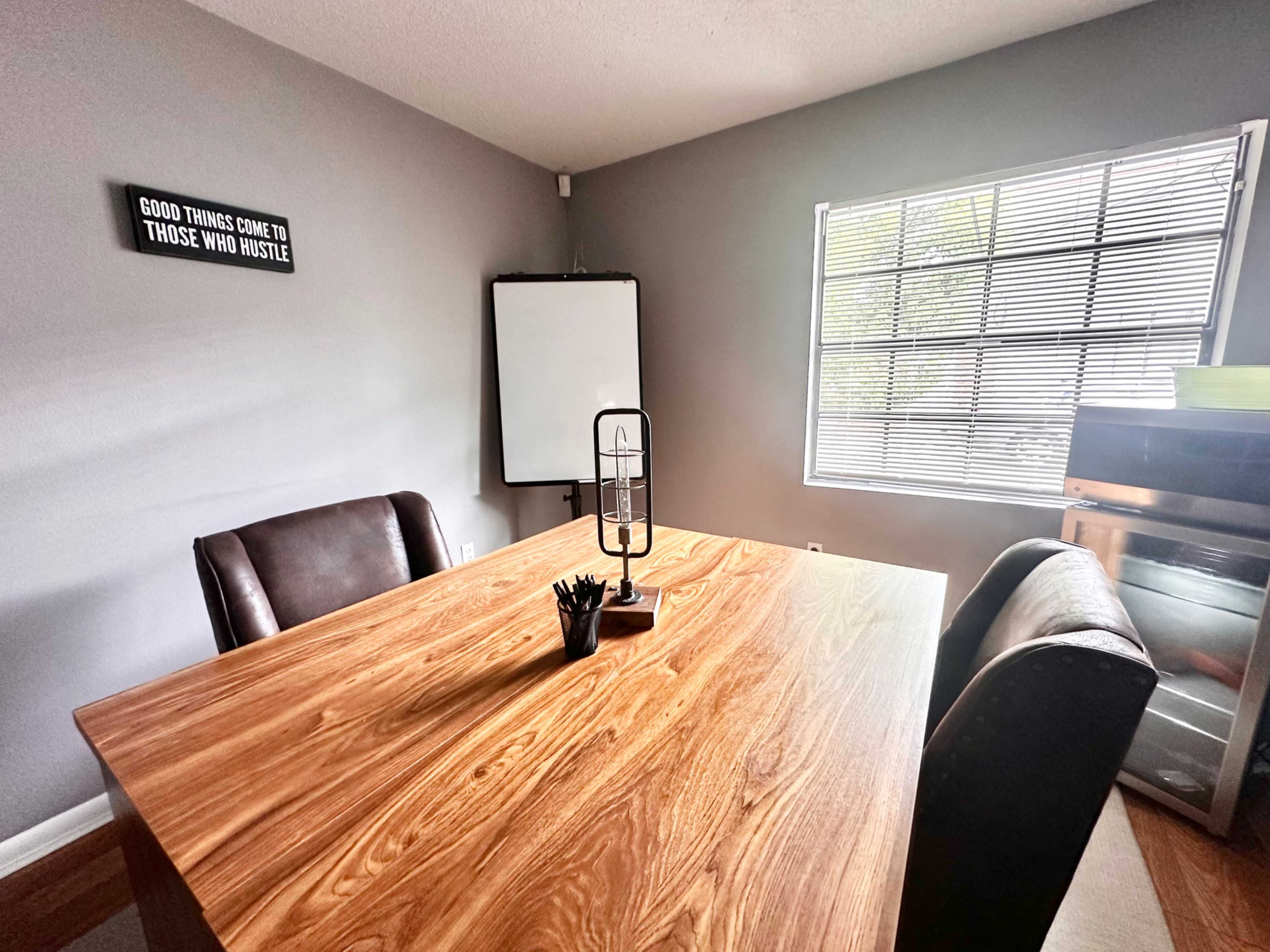 A modern workspace features a wooden table with two leather chairs, a lamp, and a whiteboard against a gray wall.
