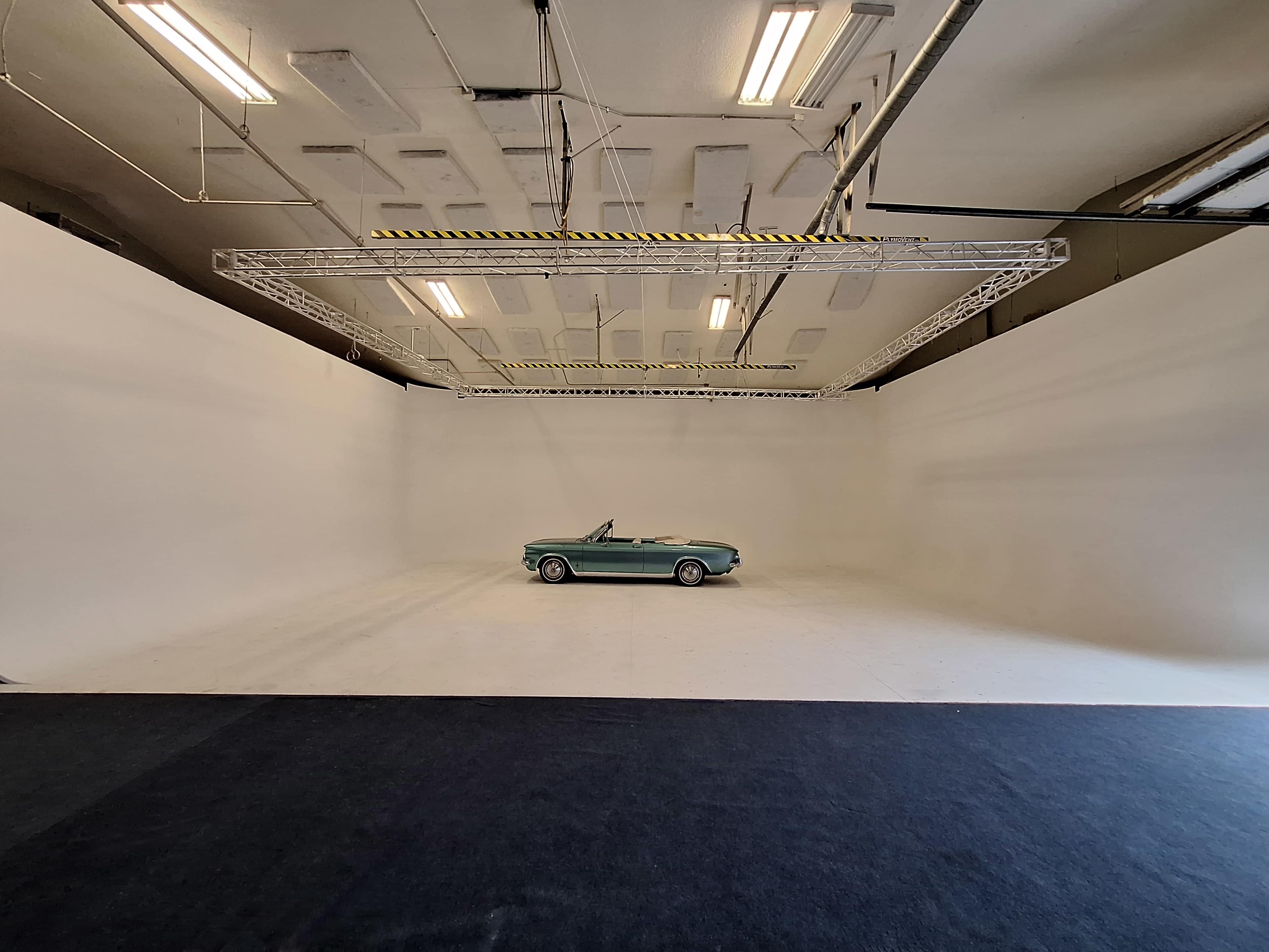 A vintage convertible car is positioned in a spacious, empty studio with white walls and overhead lighting.