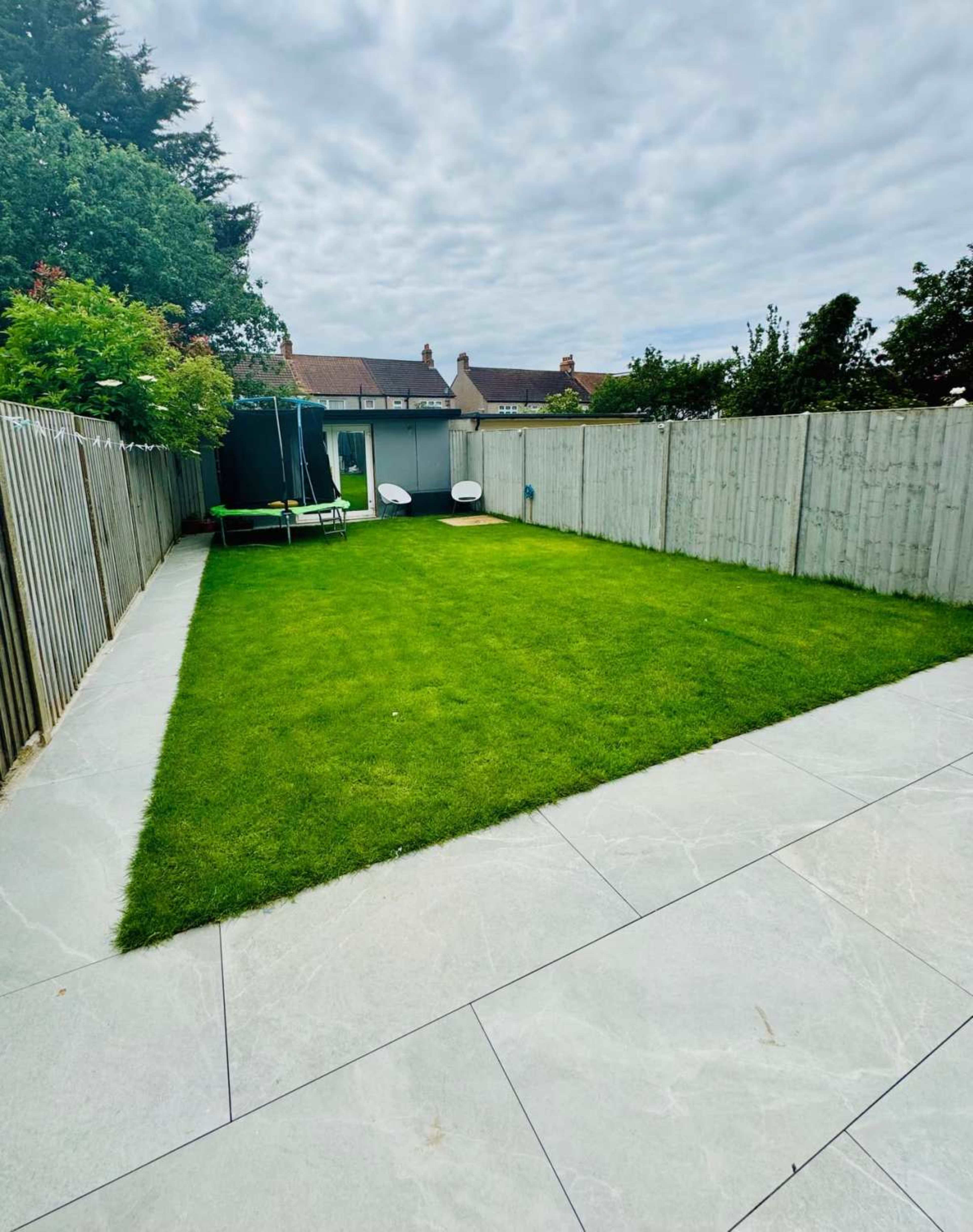 A neatly maintained backyard featuring a green lawn, a paved area, and two lounge chairs along with a swing set in the corner.